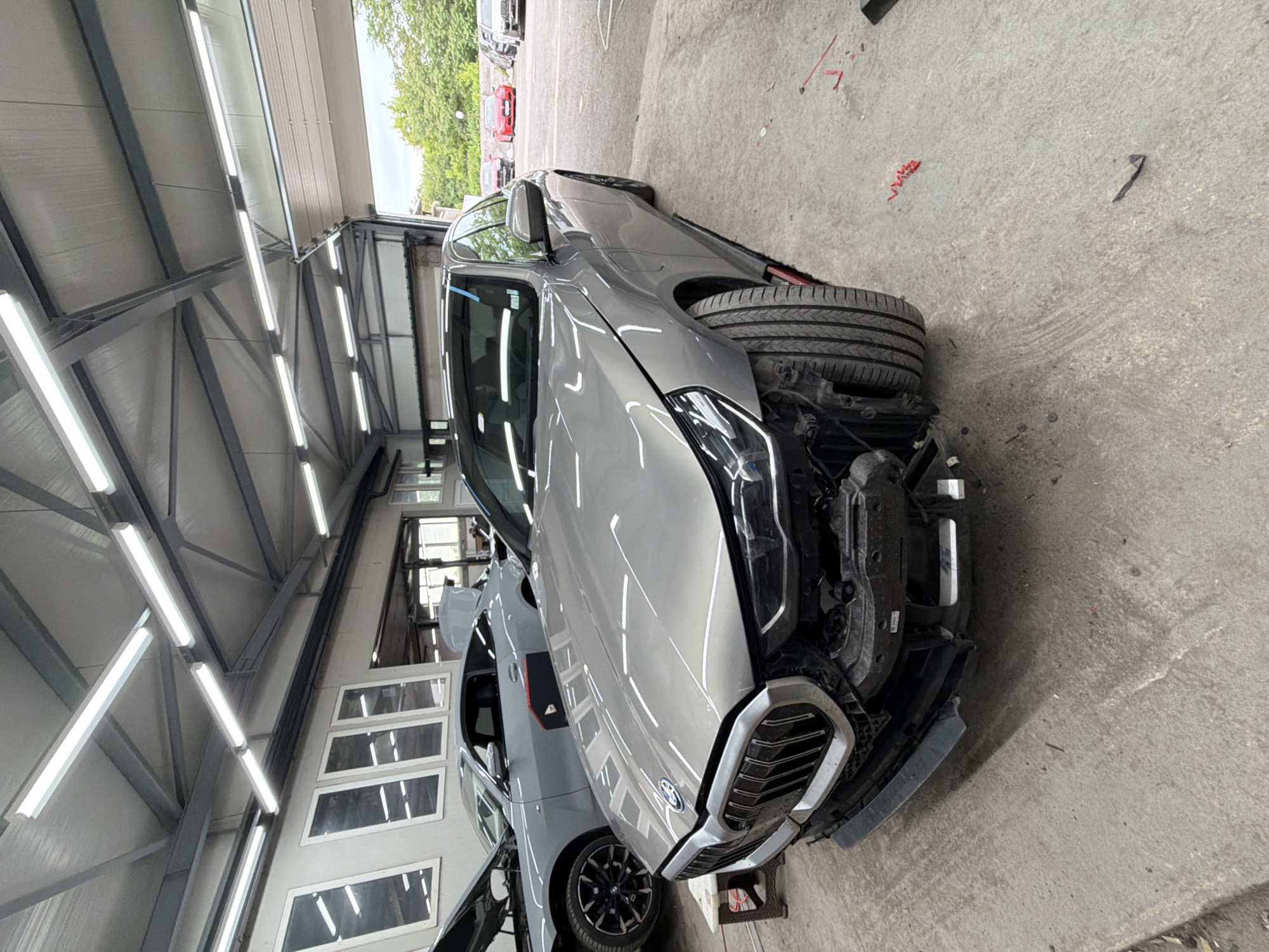 The image depicts a gray BMW car in a repair garage. The car's front bumper is removed, revealing the underlying mechanical components. Another car, also gray, is visible in the background with its doors open. The garage has a concrete floor and a high ceiling with fluorescent lighting. Through the open garage door, other vehicles and greenery can be seen, suggesting an outdoor setting. The image was taken indoors. The location is presumed to be in Burgas, Bulgaria based on the information provided. No people are directly visible in the frame. Other objects present include tools, parts of the car, and the structure of the garage itself. The car is under repair.