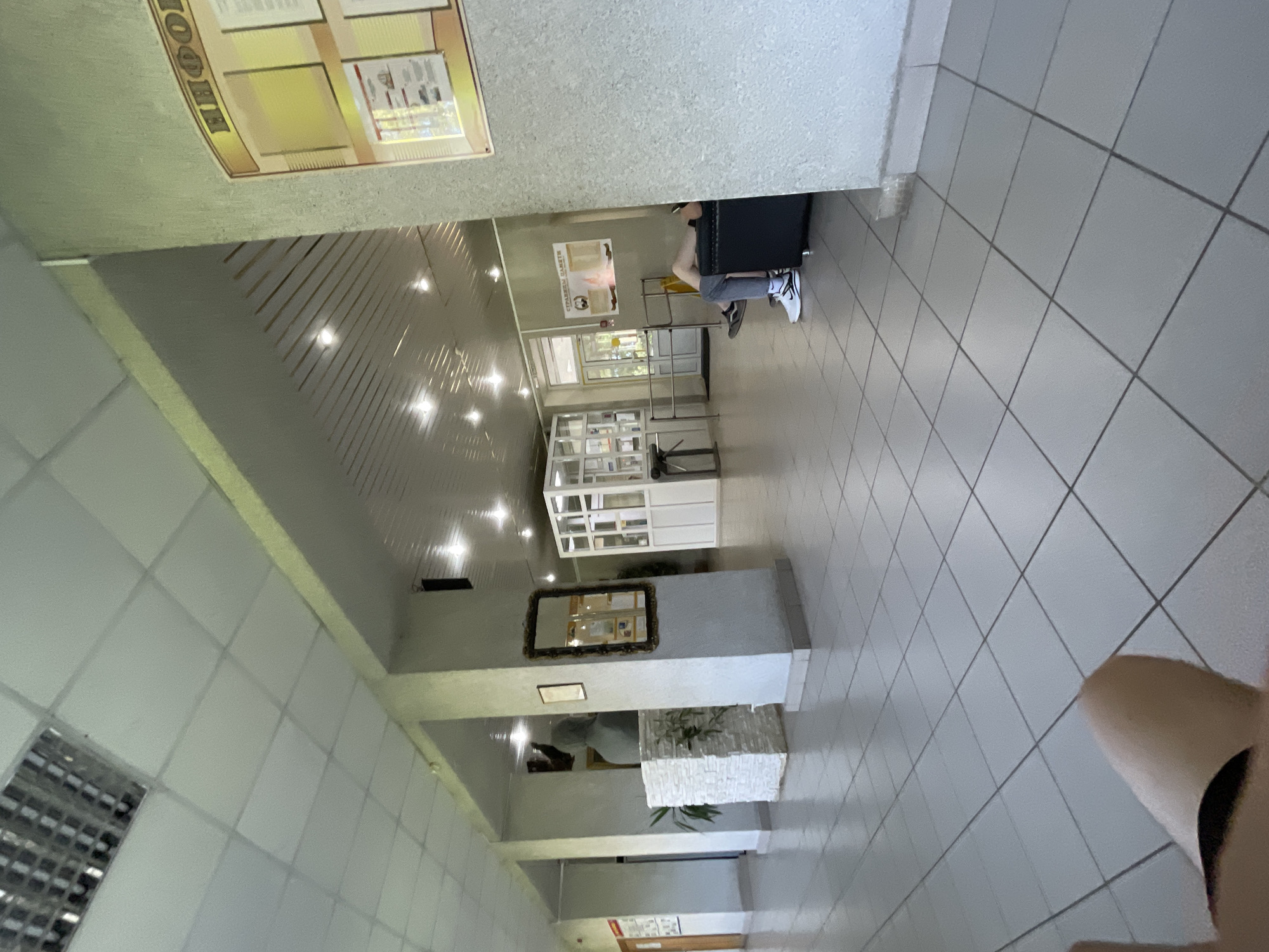 The image shows an interior view of a hallway or lobby. The floor is tiled with large, light-colored square tiles. The walls are painted a neutral color, and there are several pillars supporting the ceiling. The ceiling has fluorescent lighting fixtures and a visible suspended grid.
On the left side of the image, there is a statue bust on a pedestal adorned with white textured panels and some greenery. A framed picture hangs on the wall near the statue. Further down the hallway, there's a door and a bulletin board on the wall.
In the middle of the hallway, a glass-enclosed structure is visible, possibly a reception desk or a display area, with white framing.
On the right side of the image, a person is seated on a dark-colored sofa, facing away from the camera. They are wearing grey pants and white sneakers. To the right of the sofa, a portion of a wall is visible, with a framed bulletin board mounted on it displaying information. Above the bulletin board, text in Cyrillic letters is visible, which appears to be a heading like "ИНФО" or "ИНФОРМАЦИЯ".
The overall environment suggests a public building, perhaps an administrative office, educational institution, or a community center in Красные Баки, Russia. The lighting is bright, indicating it is daytime. The scene is static, with no apparent activity other than the seated individual.