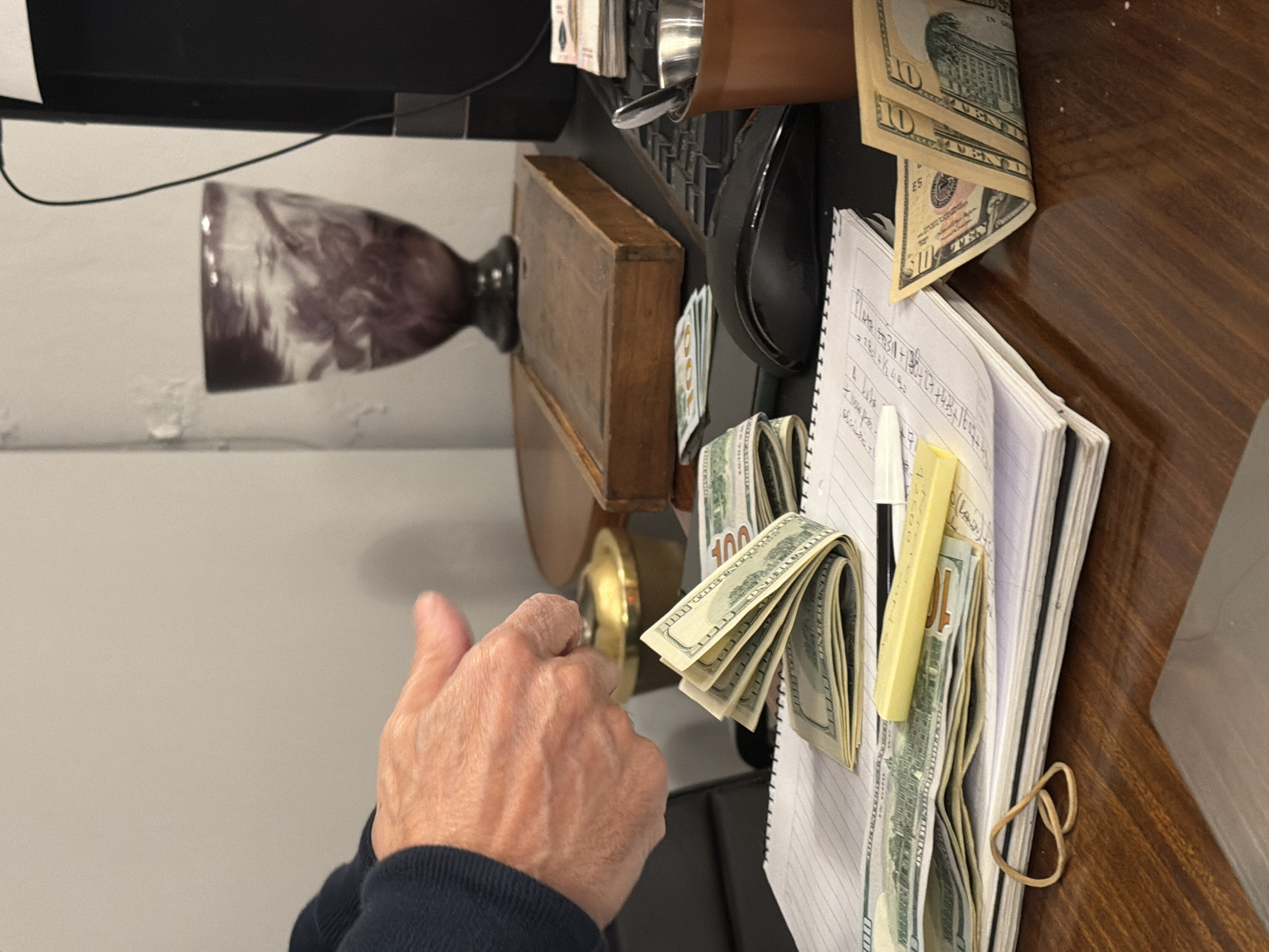 A close-up shot depicts a person turning a golden-colored doorknob with their right hand. Situated on a dark-colored desk are a computer keyboard and mouse, notebooks, money, and a pen. The money consists of a mix of $100 and $10 bills. The overall scene suggests a desk or counter area, but there are no definitive indicators of a specific location in Buenos Aires, Argentina. The wall is off-white.