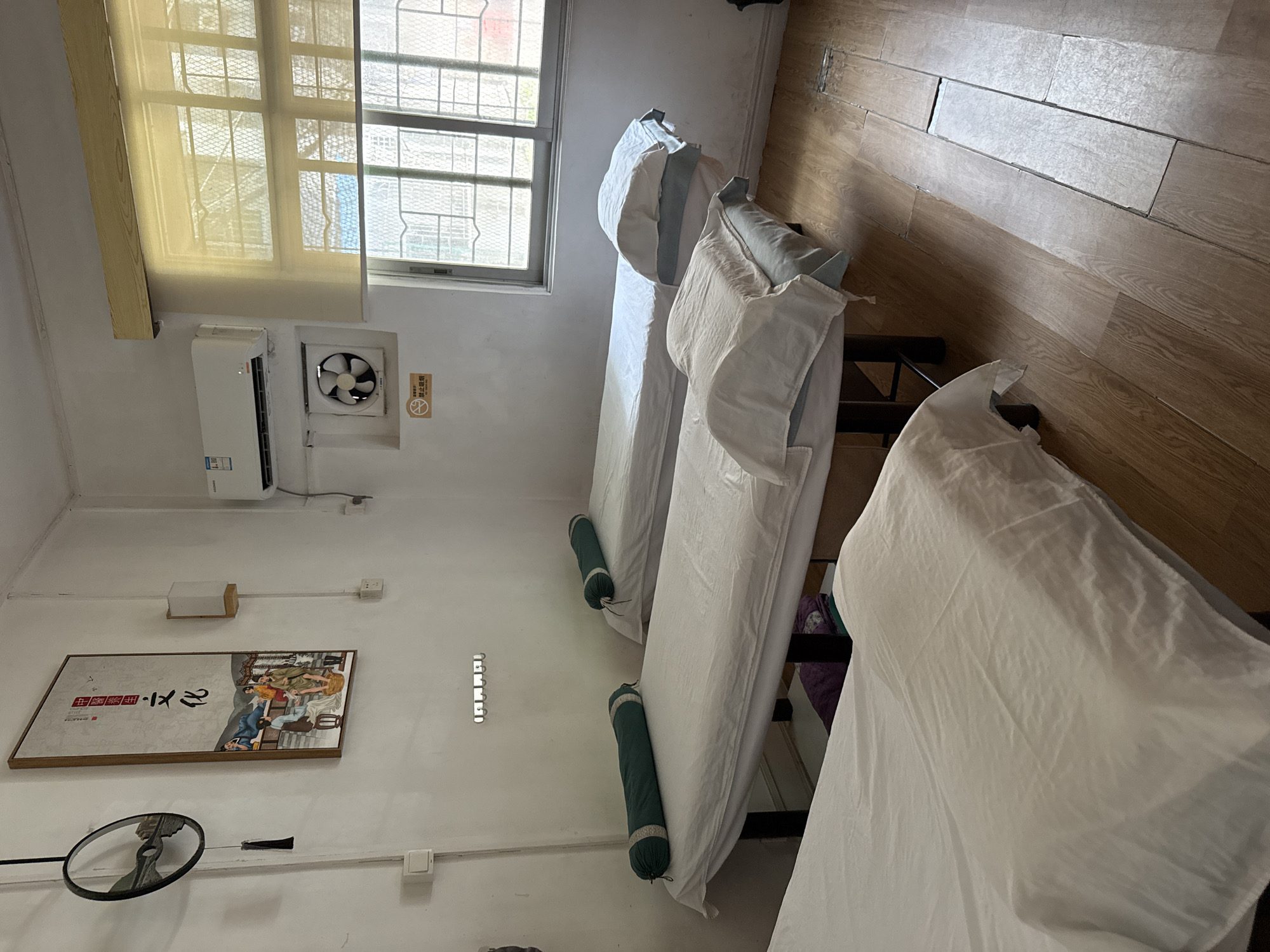 The image depicts an indoor massage or treatment room, possibly in Huizhou, China, featuring three massage beds. The beds are covered with white linens, and the two visible beds in the foreground and middle ground have green bolster pillows placed on them. The room has white walls, and a wooden floor. On the left wall, a circular decorative item with a tassel hangs, and above it is a framed artwork depicting figures in traditional Chinese attire. A modern air conditioner is mounted on the upper right wall, near a window covered with a translucent blind. Below the air conditioner, a small exhaust fan is visible in a wall recess, accompanied by a "No Smoking" sign. The overall impression is of a functional, albeit somewhat sparsely decorated, treatment space.