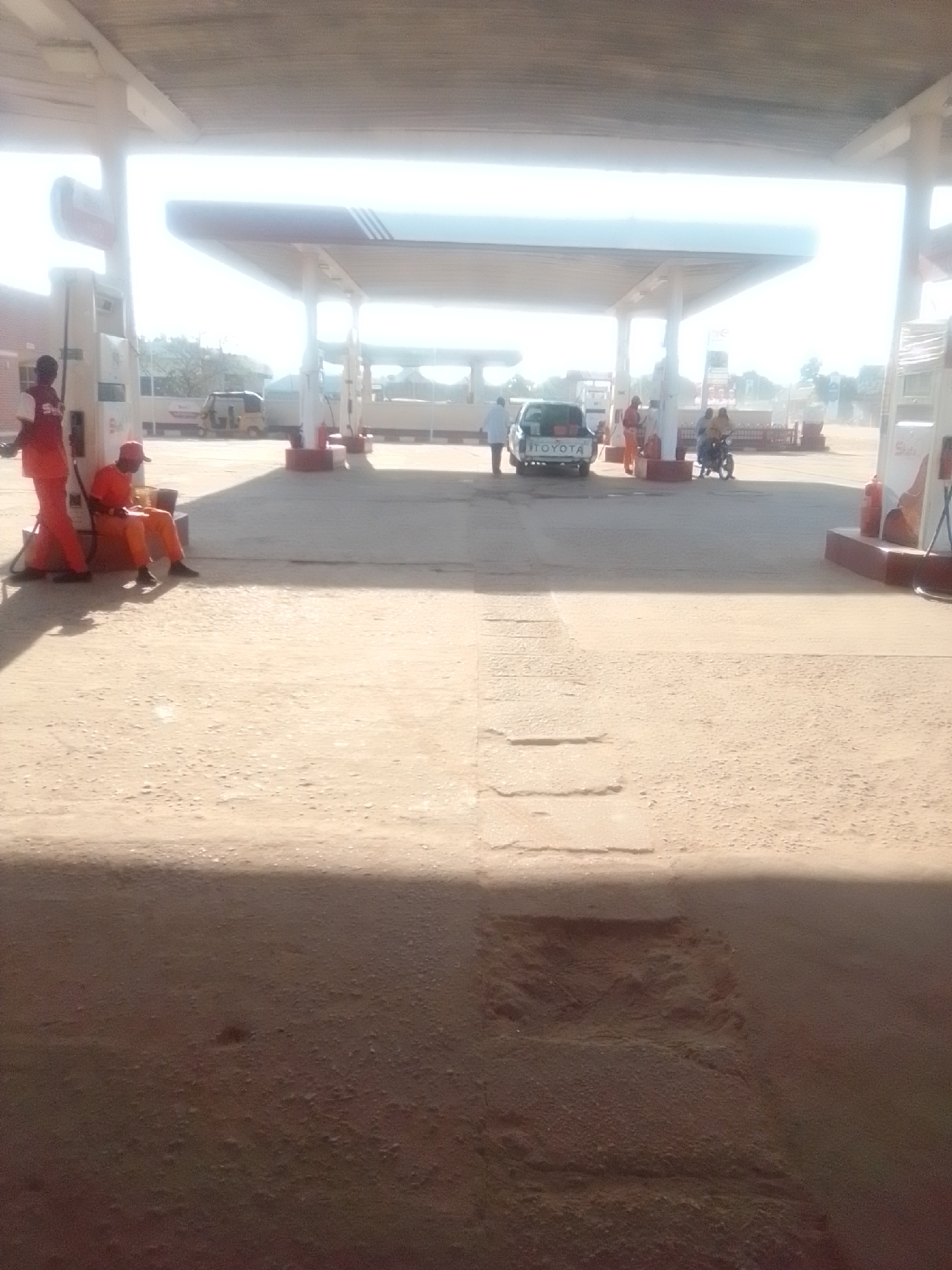 The image depicts an outdoor scene at a gas station, likely in Kwadon, Nigeria, as indicated by the context. The foreground is dominated by a sandy, uneven surface, casting a shadow from an unseen structure. In the mid-ground, under a covered canopy supported by columns, a white Toyota pickup truck is parked. To the left of the truck, a person in orange overalls and a red cap is seated, looking down at something in their lap. Another person in similar orange attire stands to the left of a fuel pump. Further to the right, two individuals are visible on or near a motorcycle. The gas station appears to be operational, with fuel pumps and the characteristic canopy structure. The lighting suggests it is daytime, with bright sunlight creating strong contrasts and some overexposure in the upper portion of the image. The environment is dusty and arid.