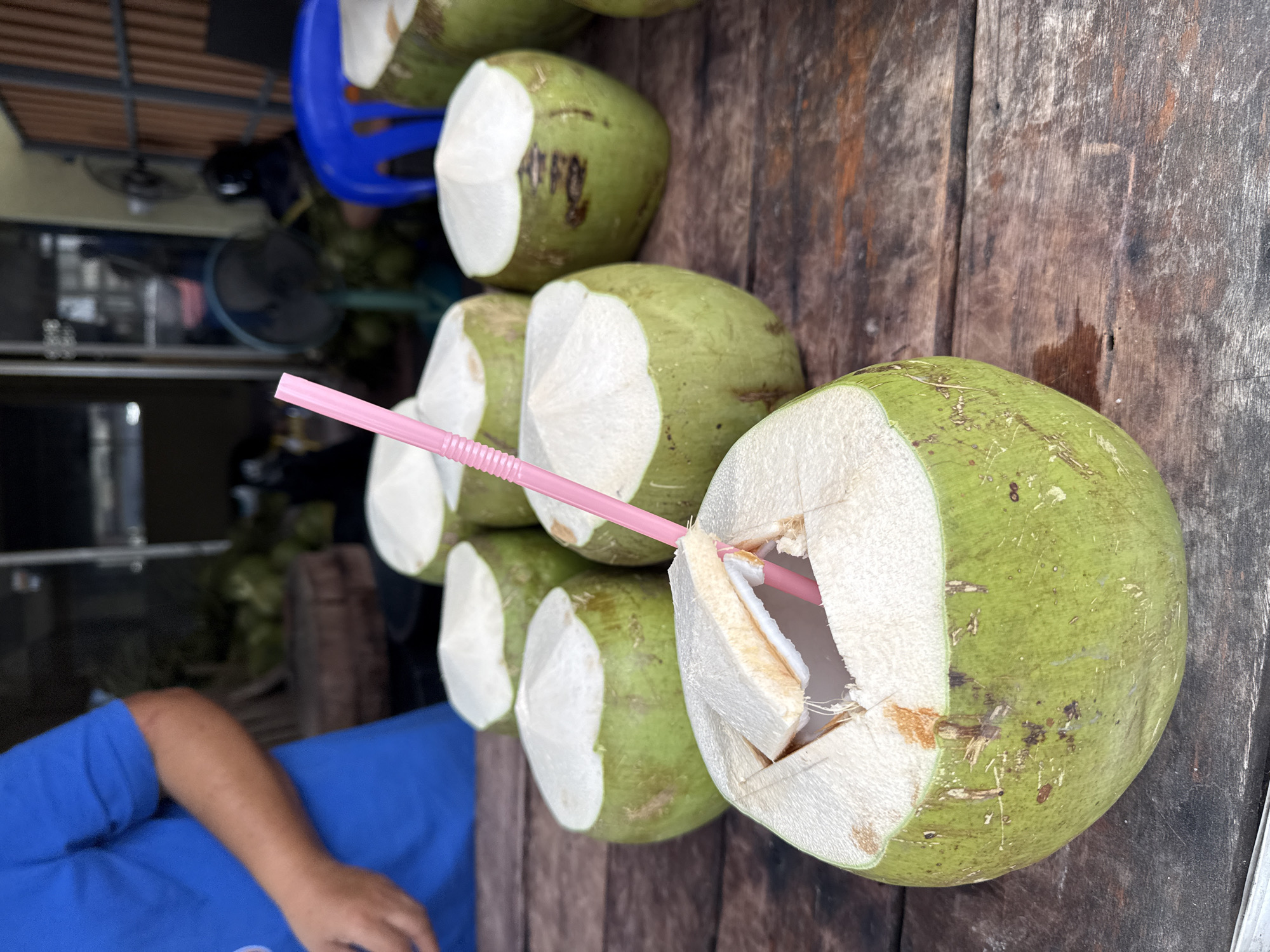 In Ko Samui District, Thailand, a close-up shot captures a display of freshly prepared coconuts on a weathered wooden table. In the foreground, one coconut is prominently featured, its top expertly cut to reveal the white flesh and water within. A pink plastic straw is inserted into this coconut, ready for consumption. Behind it, several more coconuts are arranged, their green exteriors contrasting with their white interiors. In the blurred background, a person wearing a blue shirt is partially visible, suggesting a vendor or someone preparing the coconuts. Other elements in the background include what appear to be more coconuts, a blue chair, and a fan, hinting at an outdoor market or roadside stall. The overall scene conveys a refreshing and tropical atmosphere.