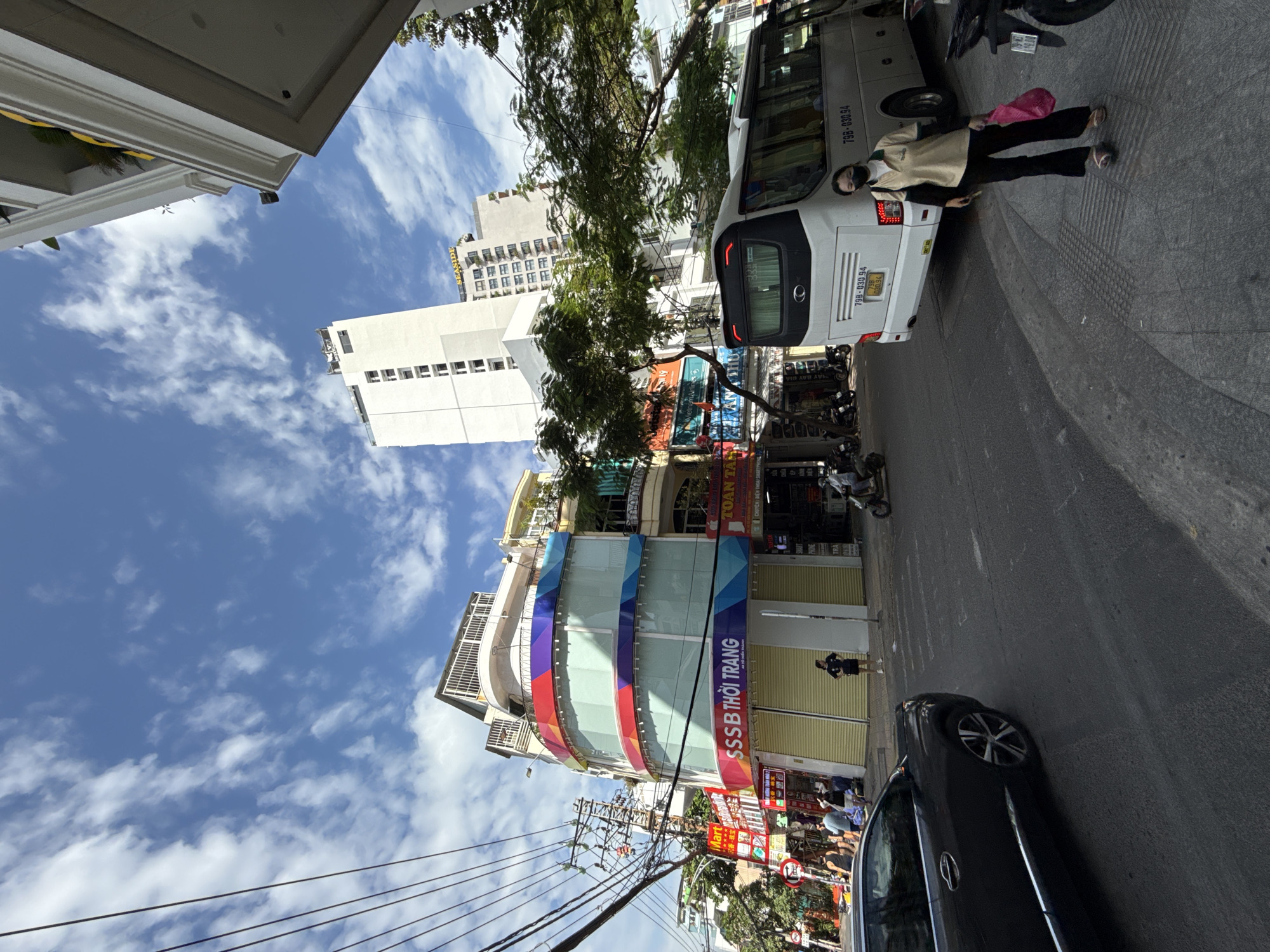 The image depicts a street scene in Nha Trang, Vietnam, on a sunny day with scattered clouds. In the foreground, a dark-colored sedan is parked on the left side of the asphalt road. The road has pedestrian crossing lines visible. On the right side of the road, a white bus with license plate 79B-030.94 is parked. A woman wearing a light-colored polo shirt, dark pants, and a black face mask is walking on the sidewalk next to the bus, carrying a pink shopping bag.
Behind the bus, a multi-story building with a curved facade is prominent, featuring windows and colorful striped decorations. The ground floor of this building appears to be a shop with yellow roller shutters, and a sign reads "SSSS THỜI TRANG." Adjacent to it, another shop sign reads "TOÀN THẮNG." Further in the background, taller white buildings, including one with a sign that says "SEASIDE," dominate the skyline. Several trees with green foliage are interspersed between the buildings. Overhead, a complex network of power lines is visible against the blue sky. Several other vehicles, including motorcycles, are parked or in motion in the background, suggesting a busy urban environment. People are seen walking on the sidewalks and near the shops.