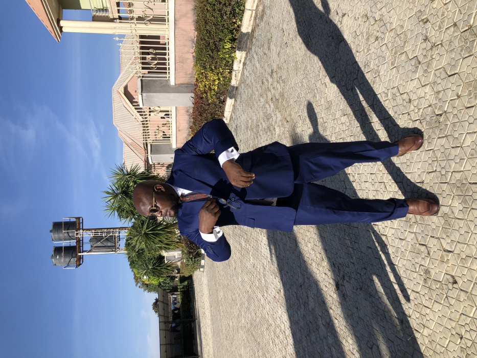 A bald man wearing a blue suit, white shirt, and brown shoes stands on a tiled surface. He has sunglasses on and appears to be adjusting his tie. His shadow stretches out on the ground. In the background, there is a building, vegetation, and a water tank structure under a blue sky. The photo appears to be taken on a sunny day.
