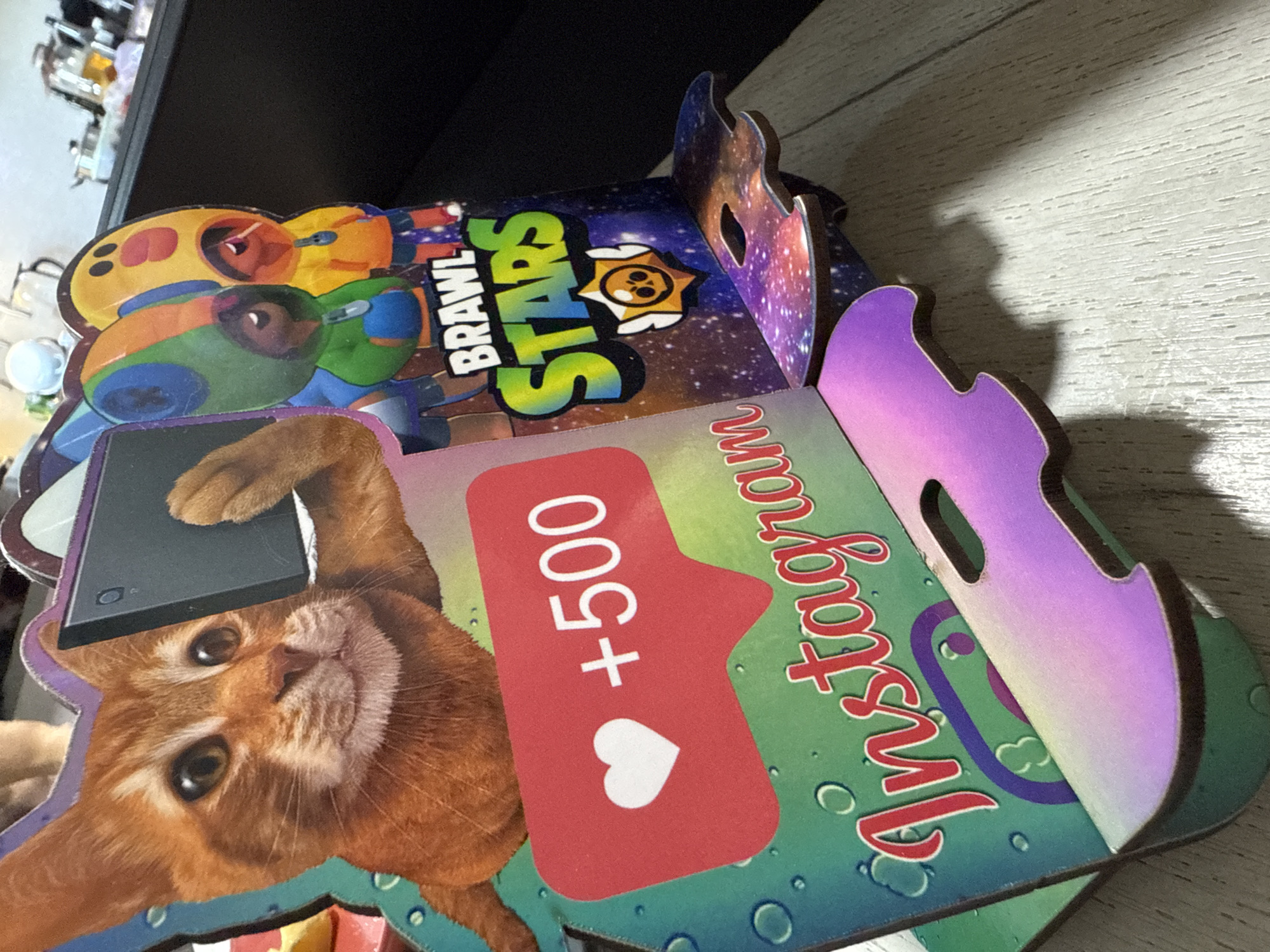 This is a close-up, eye-level shot of a brightly colored, pop-up decoration or display featuring cartoon characters and social media elements. The primary subject is a stylized orange tabby cat holding a smartphone, appearing to take a selfie. To the right of the cat are two characters from the video game "Brawl Stars," one in a green and blue hooded outfit and the other in a yellow hooded outfit. Below these characters, the "Brawl Stars" logo is prominently displayed with a star emblem. In the foreground, a red speech bubble icon with a white heart and "+500" suggests a social media interaction, likely representing likes on Instagram. The words "Instagram" are written in a stylized script across the lower part of the display, with the Instagram logo faintly visible behind it. The decoration is made of thick cardstock or a similar material, with a glossy finish. It appears to be standing on a light-colored wooden surface, possibly a table. The background is blurred, showing indistinct objects and suggesting an indoor environment. The image is taken from an angle that highlights the layered, three-dimensional nature of the display. There is no indication of the location being in Kroyanskoye, Russia.