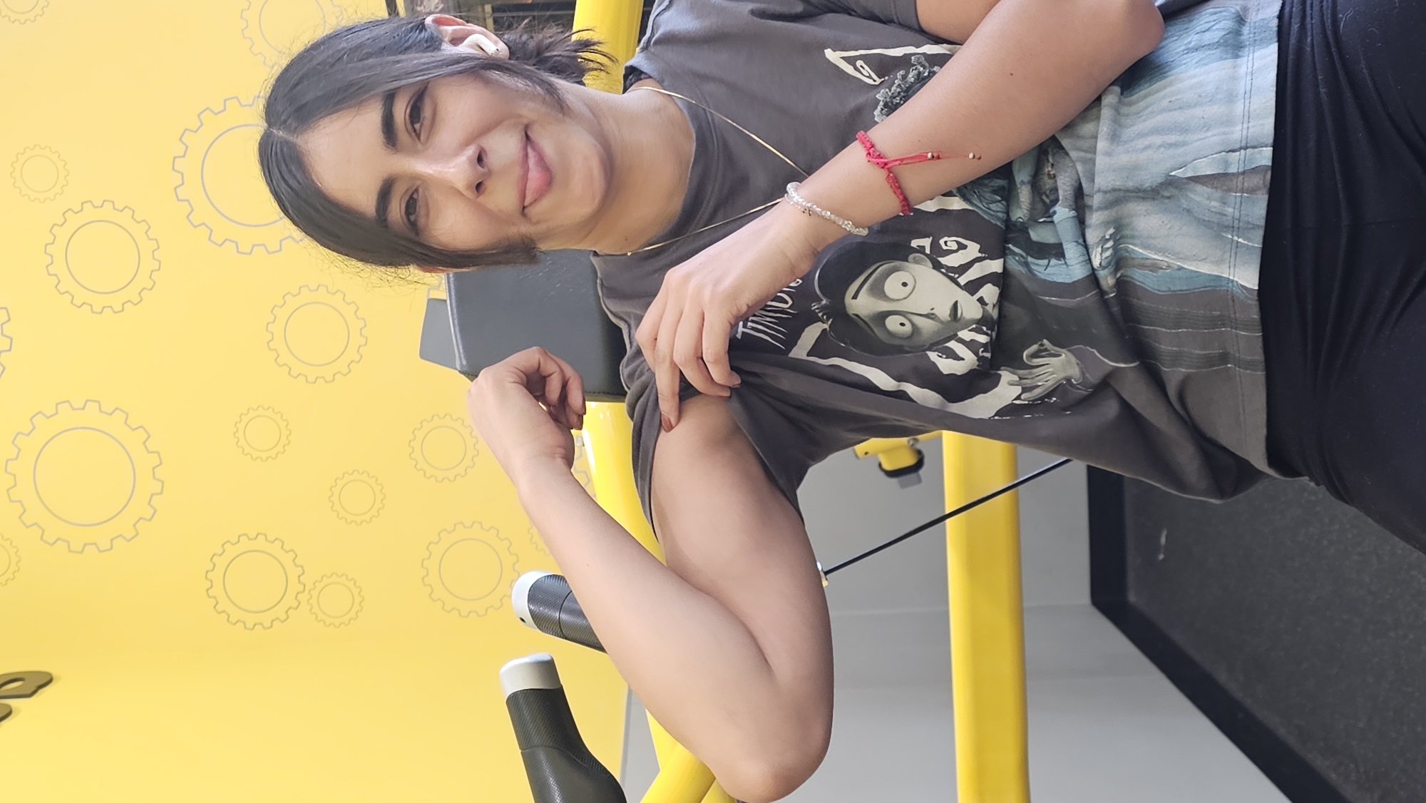 A woman is standing indoors in front of a yellow wall decorated with a pattern of gray circles. She is wearing a gray t-shirt with a graphic on it and black leggings. The woman is flexing her left bicep and smiling. The background includes yellow metal exercise equipment. It is not possible to determine the location in Puebla, Mexico from the image.