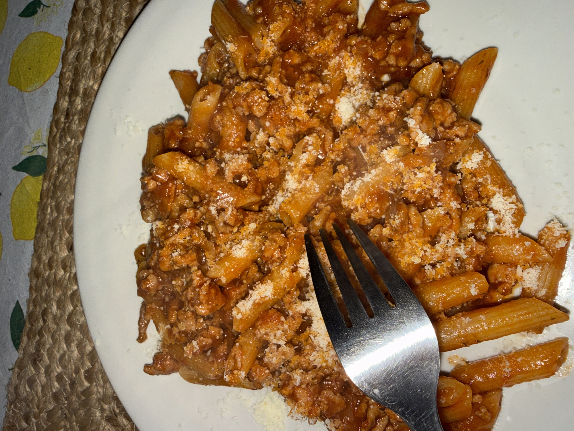 A plate of penne pasta with meat sauce and grated cheese sits on a white plate. A metal fork rests on top of the pasta. The plate is positioned on a table with a tablecloth featuring a lemon print and a placemat made of woven material. The location is likely indoors. It's not possible to confirm the location is Vozdovac Urban Municipality, Serbia without additional information.