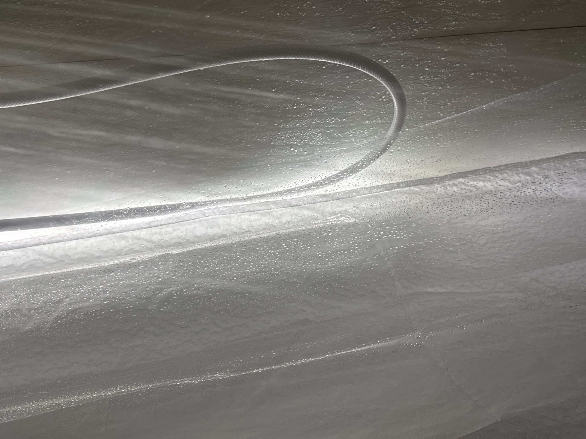 The image shows a close-up of a white shower curtain with water droplets on it, illuminated by a light source from the top left. The curtain is draped in folds, creating texture and shadows. A white shower hose is visible on the right side of the image, curving downwards. The lighting creates a slightly hazy or steamy effect. The setting appears to be a bathroom. There are no people or animals visible in the image. The image is a photograph taken from a low angle, looking up at the shower curtain and hose. The location is not specified in Saratov, Russia, and no specific details about the city can be inferred from the image.