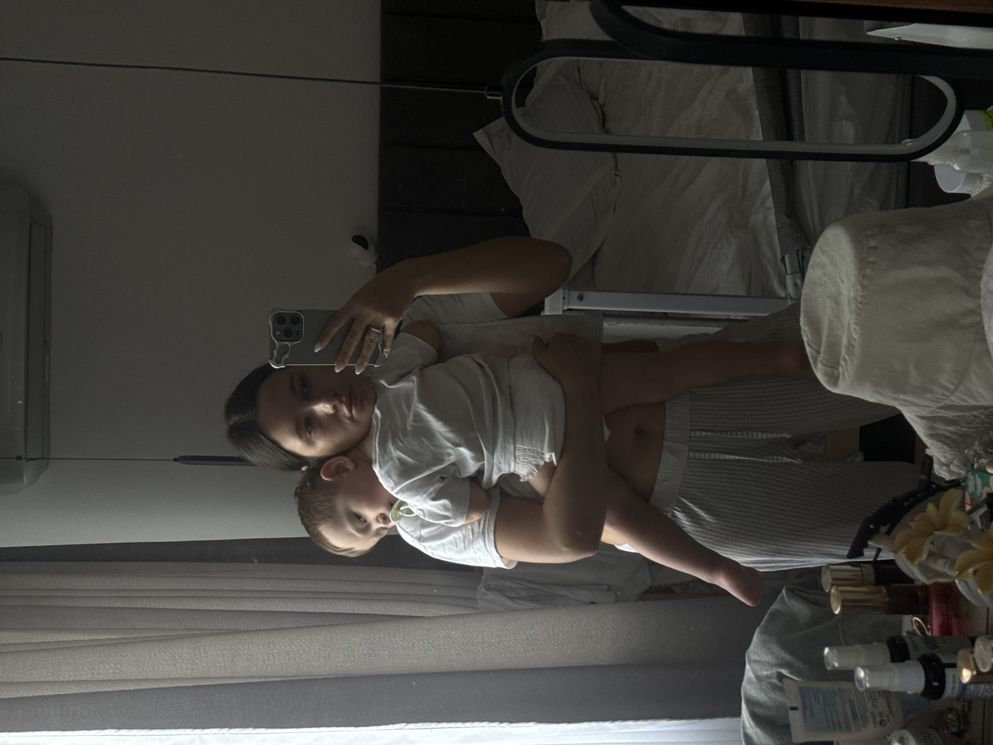 The image is a mirror selfie capturing a woman holding a young child in an indoor setting, likely a bedroom or living area. The photo appears to be taken by the woman holding a smartphone, visible in her hand, which features a multi-lens camera array and a silver-toned case.
The woman, with dark hair, is on the left side of the frame, looking directly at her reflection in the mirror (and thus at the camera). She wears a light-colored top and light-colored striped pants. In her arms, she cradles a young child, who appears to be a boy. The child has light-colored hair and a neutral expression, also looking towards the camera, with a pacifier in his mouth. He is dressed in a light-colored top.
In the background of the reflection, part of a bed with white bedding and a dark bed frame or headboard is visible on the upper right side. On the far left, a white air conditioner unit is mounted on the wall. Light-colored curtains are partially visible on the lower left of the frame. To the right of the subjects, various bottles, likely cosmetics or toiletries, are arranged on a surface, some with gold-colored caps, alongside a few decorative items. The room is moderately lit, suggesting daytime, and has a calm, domestic atmosphere. No discernible text is visible on any objects within the frame.