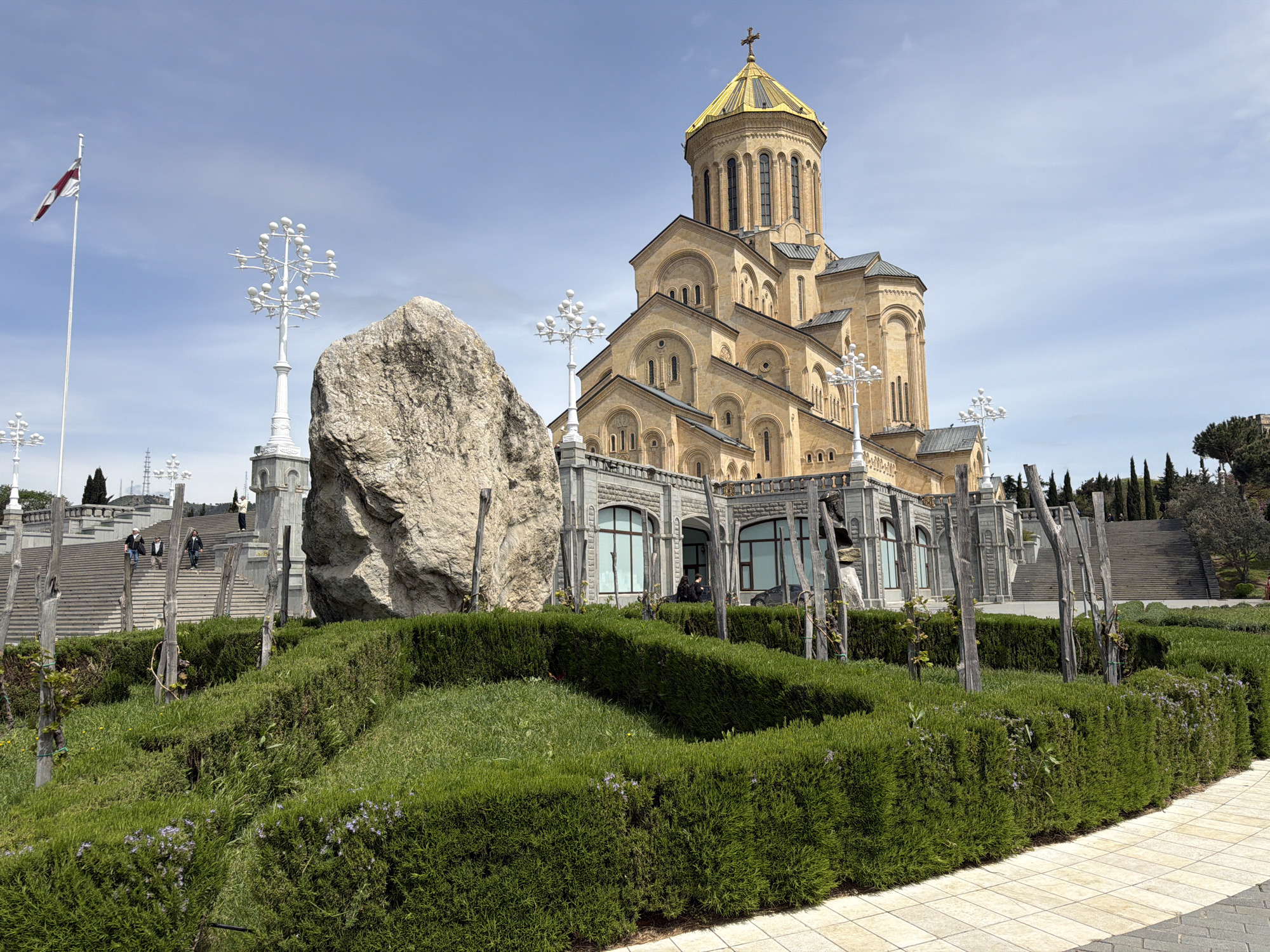 A grand Orthodox cathedral with a striking golden dome and light-colored stone facade dominates the center of the image, set against a bright blue sky with scattered white clouds. The cathedral features multiple tiered sections, arched windows, and is crowned with a cross.
In the foreground, a well-maintained garden area features manicured green hedges and grass, with a large, rough-hewn light gray boulder positioned prominently on the left. Several ornate white lampposts, resembling multi-branched structures, are scattered throughout the grounds, adding a decorative element. To the far left, a tall flagpole proudly displays the red and white flag of Georgia.
Stone steps and walkways lead up towards the cathedral, with a few people visible walking on them and near the building's base, suggesting public access to the site. The scene is captured during clear daytime under sunny conditions. In the background to the right, tall evergreen trees line the landscape. The overall atmosphere conveyed is one of grandeur and serenity, typical of a significant religious landmark. No discernible text is visible within the image.