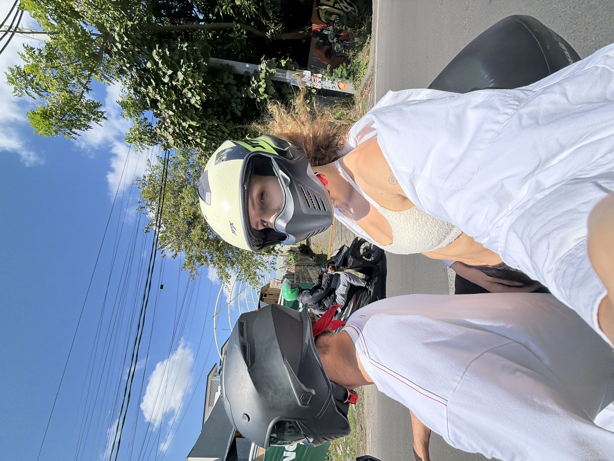 Two individuals are riding a motorcycle in North Kuta, Indonesia, on a sunny day. The person in the foreground, wearing a light-colored crop top and a white open shirt, is looking directly at the camera and wearing a yellow and black helmet with a black face mask. Their hair is styled in loose waves. The person in the background, wearing a white t-shirt and a black helmet, is not fully visible. In the distance, another person on a motorcycle is wearing a black jacket and a green helmet. The scene takes place on a paved road with trees and buildings along the sides, under a bright blue sky with scattered clouds. Overhead power lines are visible against the sky.