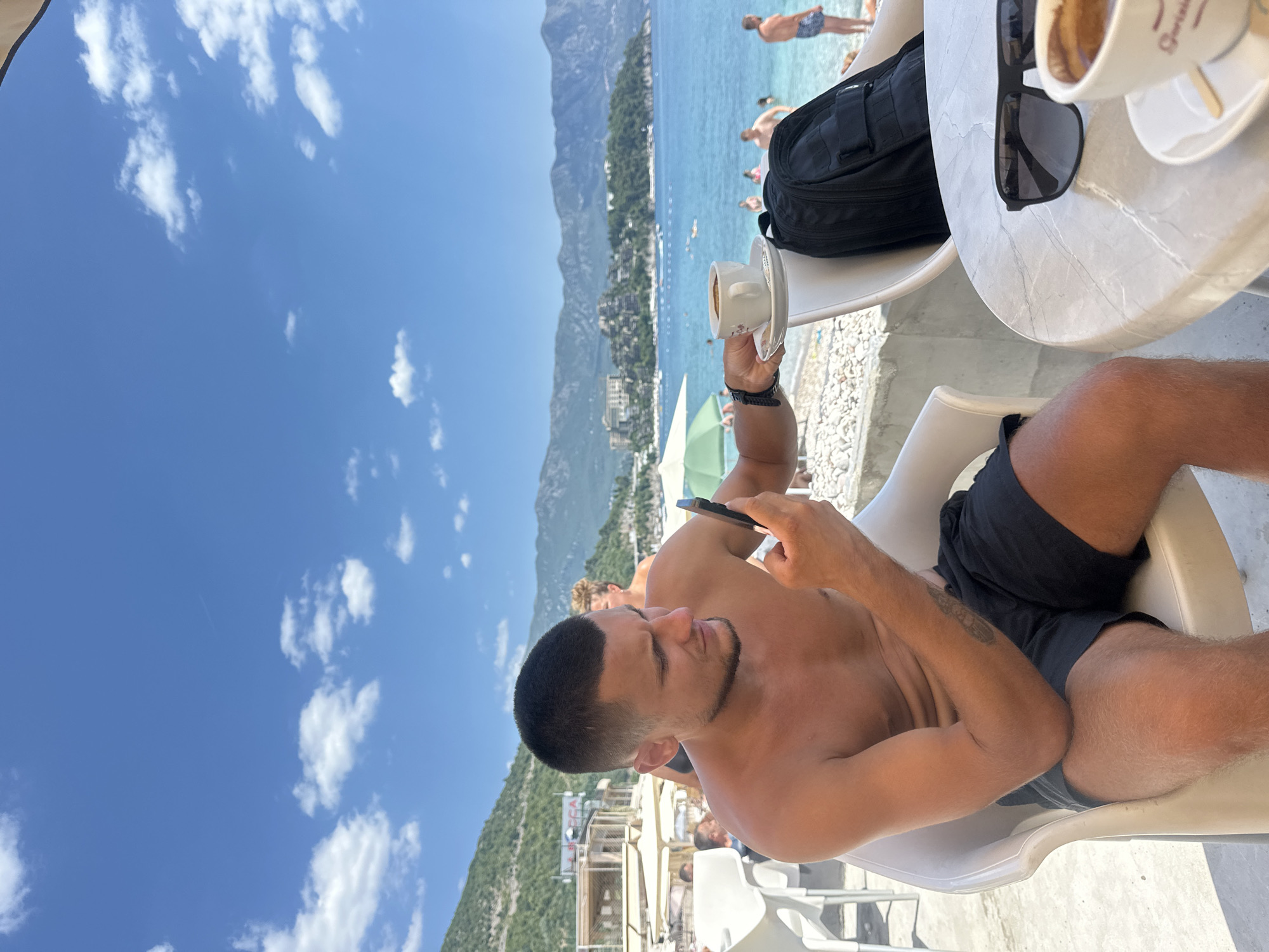 A young man with a short haircut and a beard, wearing black shorts, is sitting in a white plastic chair at a table on a pebble beach. He is shirtless, exposing a tattoo on his left arm. He holds a smartphone in his right hand and looks at it. On the table, there is a cup of coffee with a saucer, sunglasses, and a black backpack. The background shows the Adriatic Sea, with several people swimming and sunbathing. Mountains covered with vegetation rise in the distance. The sky is blue with scattered white clouds. The scene is set in Budva, Montenegro, with buildings visible on the mountainside and a resort-like structure on the beach.