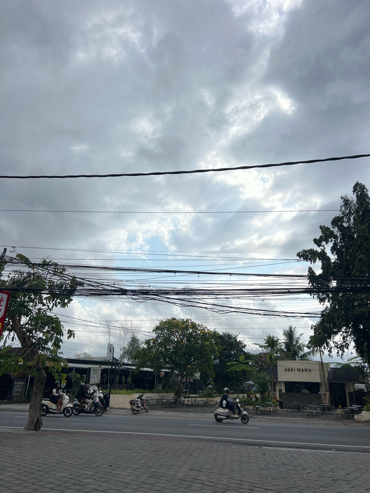 The image depicts a street scene in Denpasar, Indonesia, on a cloudy day. The foreground shows a paved sidewalk next to a road. Several motorbikes are visible on the road; one is carrying two people, another has a single rider, and a third is parked. Across the road, there are buildings with signs, including one that reads "KOPI MARU." Trees and other vegetation line the street, and a dense network of overhead electrical wires crisscrosses the sky. The overall atmosphere is overcast and muted.