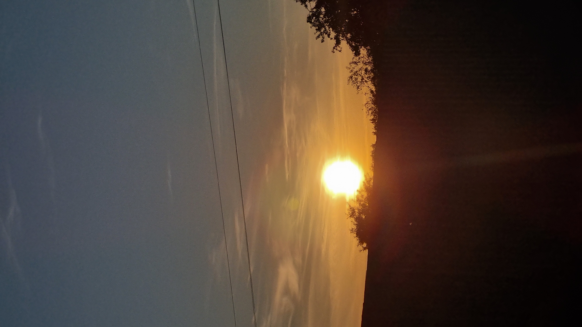This is a vertical, eye-level, medium shot of a sunset in Vysokoe, Russia. The sun is a bright white orb in the center of the frame, casting a golden-orange glow on the sky and clouds. The horizon is silhouetted by dark trees and a darker landscape. Two thin power lines are visible across the sky. The sky transitions from a pale blue at the top to a warmer orange near the horizon. There are no people visible in the image.