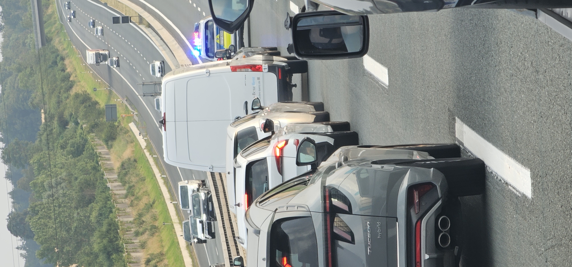 The image captures a traffic jam on a multi-lane highway. In the foreground, a grey Hyundai Tucson Hybrid is visible from the rear, stopped in traffic. To its right, the rear of a white SUV with illuminated taillights is also stopped. Further ahead and to the right, a white Mercedes-Benz Sprinter van is parked, with a police vehicle displaying flashing blue lights immediately behind it. The police vehicle has license plate "BTZP 8058". The highway is lined with trees and vegetation on both sides, with a sound barrier visible to the left. A bridge crosses the highway in the distance. The scene is consistent with traffic congestion on a German autobahn.