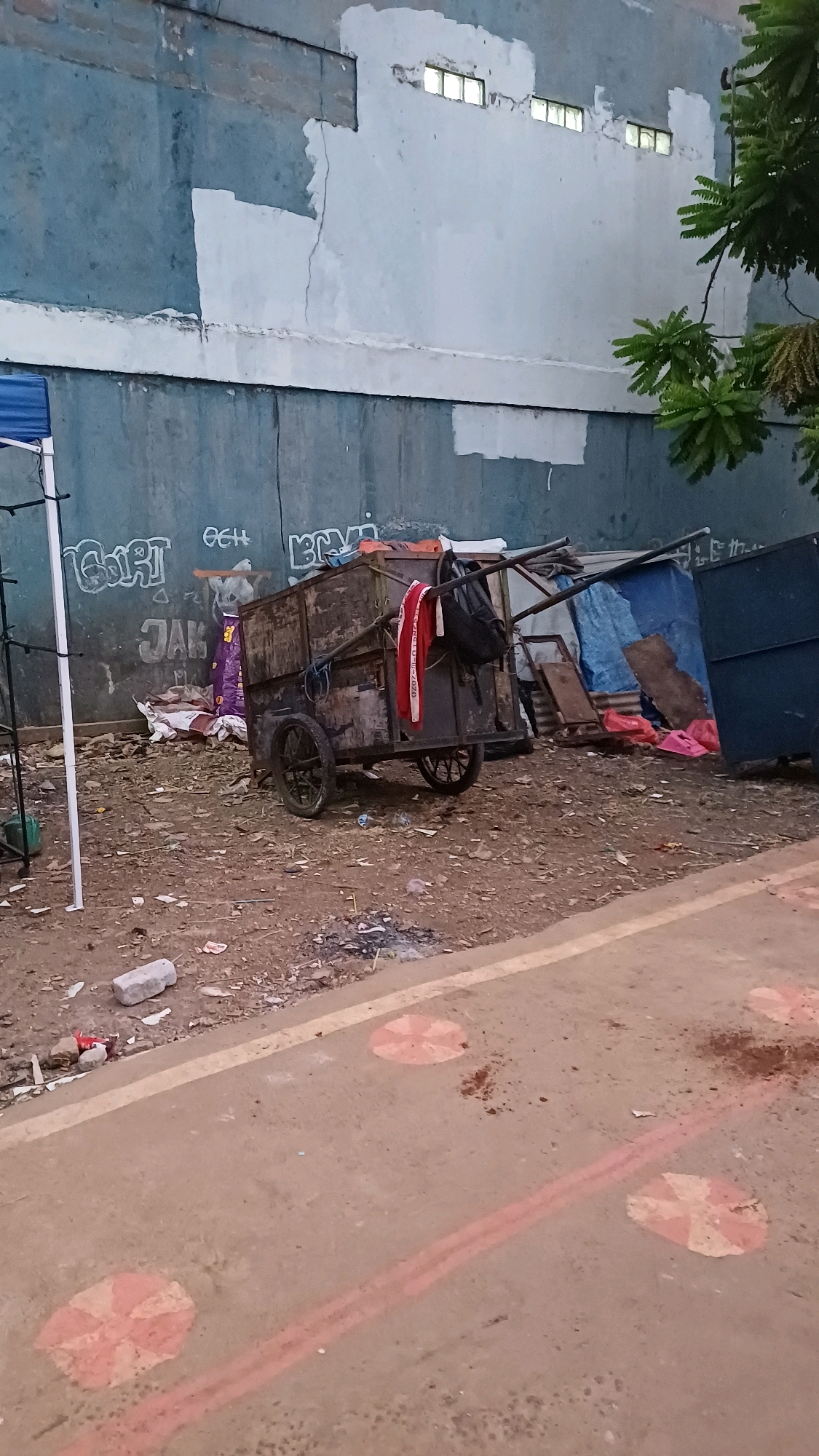 An outdoor, ground-level, medium shot depicts a weathered, two-wheeled cart laden with assorted items against a concrete wall in Cibinong, Indonesia. The cart, constructed of wood and metal, appears to be used for hauling goods or as makeshift shelter. It is positioned on a dirt and debris-strewn area, with the wall behind it covered in graffiti. A blue tarp is visible draped over the cart's frame, suggesting it may serve as a temporary shelter. Red and white fabric hangs from the cart, along with a black backpack. To the left, a white pole stands, possibly part of a larger structure. In the foreground, a paved surface with red markings and circular patterns runs parallel to the scene. The overall environment suggests a neglected urban space. There are no people visible in the image.