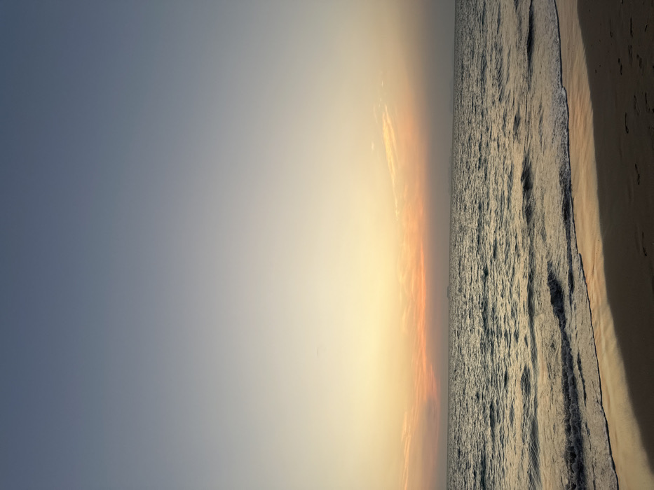 The scene displays a sunset view across a body of water from a sandy beach at Phra Ae, Thailand. The sky presents a vertical gradient, starting with deep blue at the top, transitioning to lighter blue, then pale yellow, and concluding with a horizontal band of orange and pink at the horizon. Several wispy clouds are visible within the orange-pink portion of the sky. The sea surface is dark and moderately textured with small waves, reflecting the warm colors of the sky. A strip of wet, light brown sand occupies the immediate foreground, showing reflections of the sky and faint human footprints. On the distant horizon, a landmass or island chain is visible as a hazy, dark silhouette towards the right. A single large, dark vessel or ship is discernible on the far left horizon. No individuals or explicit actions are present within the frame.