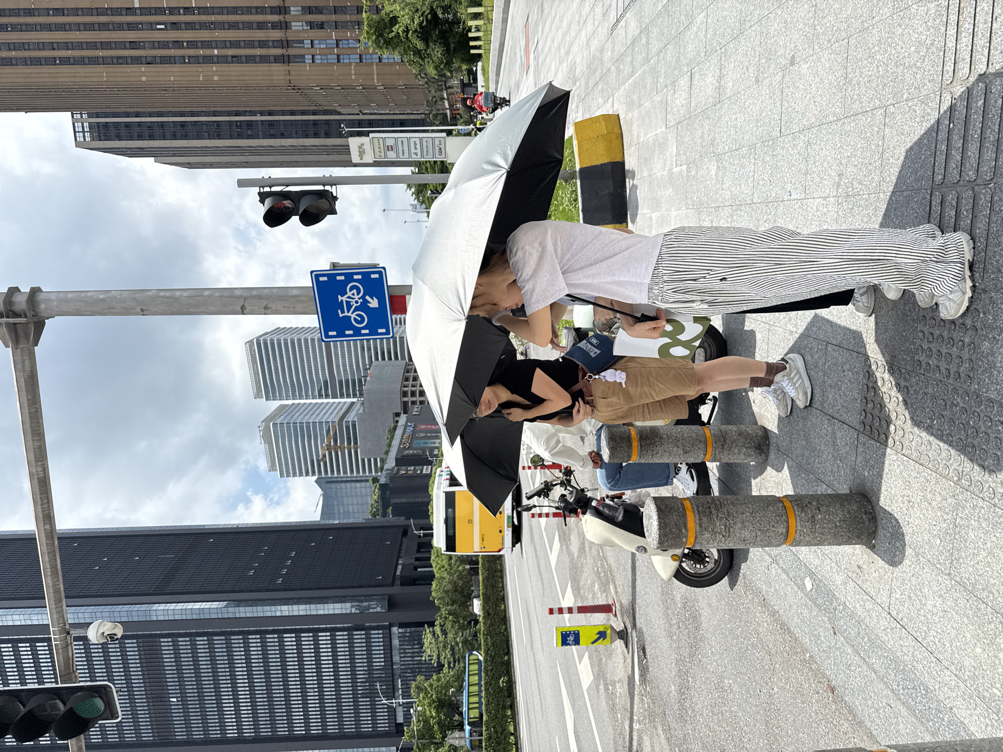 In Haizhu District, China, two individuals are standing on a paved sidewalk under a large silver umbrella, seemingly waiting. A third person is partially visible seated on a scooter to their left. The background features modern, tall buildings, a busy street with a yellow bus and a motorbike, and a traffic light. The weather appears partly cloudy.