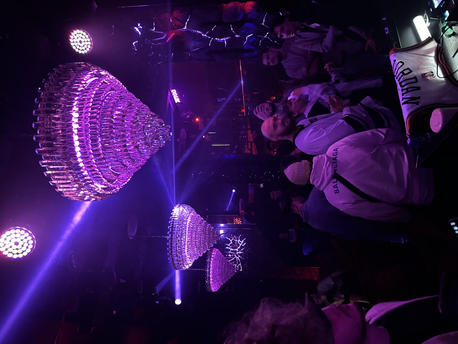 The image depicts an indoor scene within the ZOO Club, Moscow, Russia, characterized by strong purple ambient lighting.
The environment is a dark, enclosed space with multiple sources of purple illumination. Overhead, three large, tiered, inverted-cone or heart-shaped chandeliers, composed of numerous reflective elements, emit a vibrant purple glow. Several circular LED spot lights with individual diodes are also mounted on the ceiling, casting distinct purple beams through the hazy air. A stylized white sign reading "3OO" (representing "ZOO") is visible on a dark background in the upper-mid right. Textured, abstract dark wall decor with intermittent blue and red highlights is present on the right side.
Approximately 10-15 individuals, predominantly male, are visible standing in the mid-ground and background, either socializing or observing their surroundings. In the central mid-ground, a bald man with a beard, wearing a white jersey-style shirt with dark stripes, faces partially left while looking towards the viewer. To his left, another man wears a light-colored hoodie with "BALENCIAGA" printed on the back and a striped beanie, facing away from the viewer. Other individuals in the mid-ground and background are dressed in casual attire. The left side of an individual's head and shoulder with wavy, light-colored hair is visible in the immediate left foreground, facing right.
In the immediate foreground, a dark surface serves as a counter, upon which a white, folded "JORDAN" sports jersey is draped. A white plastic container and a small green object are also present on this surface. No specific active interactions are discernible; the individuals appear to be in a passive social gathering.