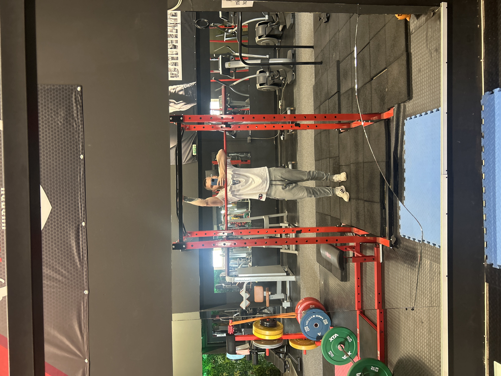 A medium shot taken indoors shows a man in a gym posing in front of a weight rack, visible in the gym's mirror. He is wearing a white t-shirt, gray sweatpants, and white sneakers. He is holding a mobile phone in his right hand and looking at the screen. The weight rack is red. Behind the weight rack, various gym machines are visible, including a seated leg press and exercise bikes. Colored weight plates are racked on a lower portion of the weight rack in the foreground. The gym has black walls, and a black banner is visible to the left. The floor is covered in a mixture of dark gray rubber tiles and blue foam tiles.