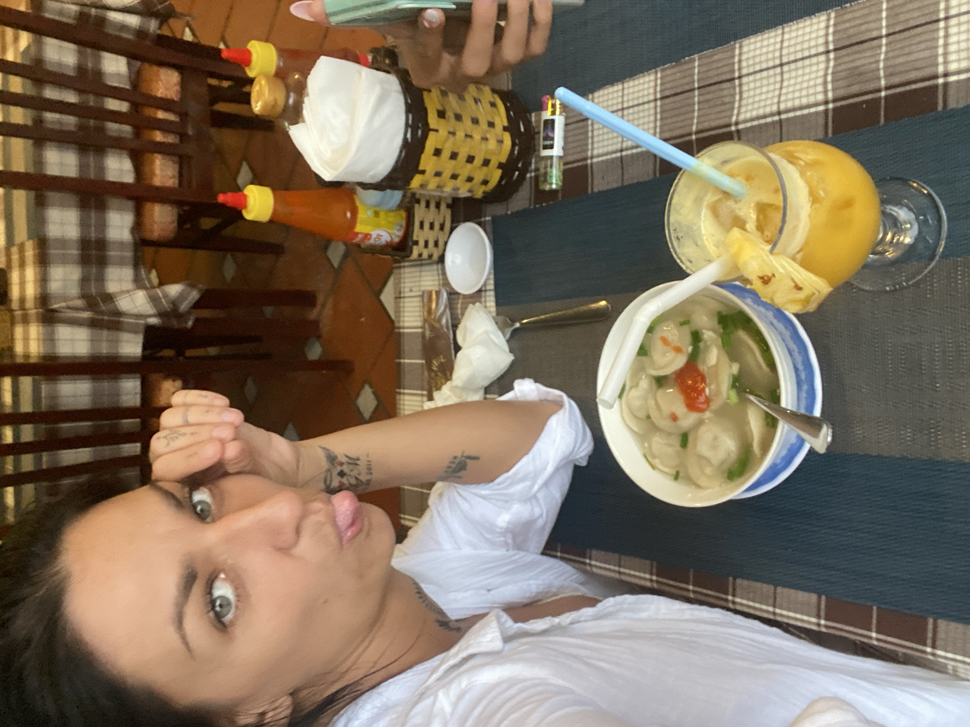 A woman is pictured in a restaurant, likely in Phu Thuy Ward, Vietnam, based on the presence of Vietnamese food items. She is wearing a white top and is positioned in the foreground, making a face for the camera. On the table are a bowl of Vietnamese soup (likely Banh Bot Loc Soup), a glass of fruit juice with a straw, two bottles of hot sauce, a basket containing napkins, a small white bowl, a lighter, a fork, and napkins. The table has a blue and brown checkered tablecloth. In the background, there is a wooden chair with a checkered cushion. Another person’s hands are visible holding a phone. The setting suggests an indoor dining environment.