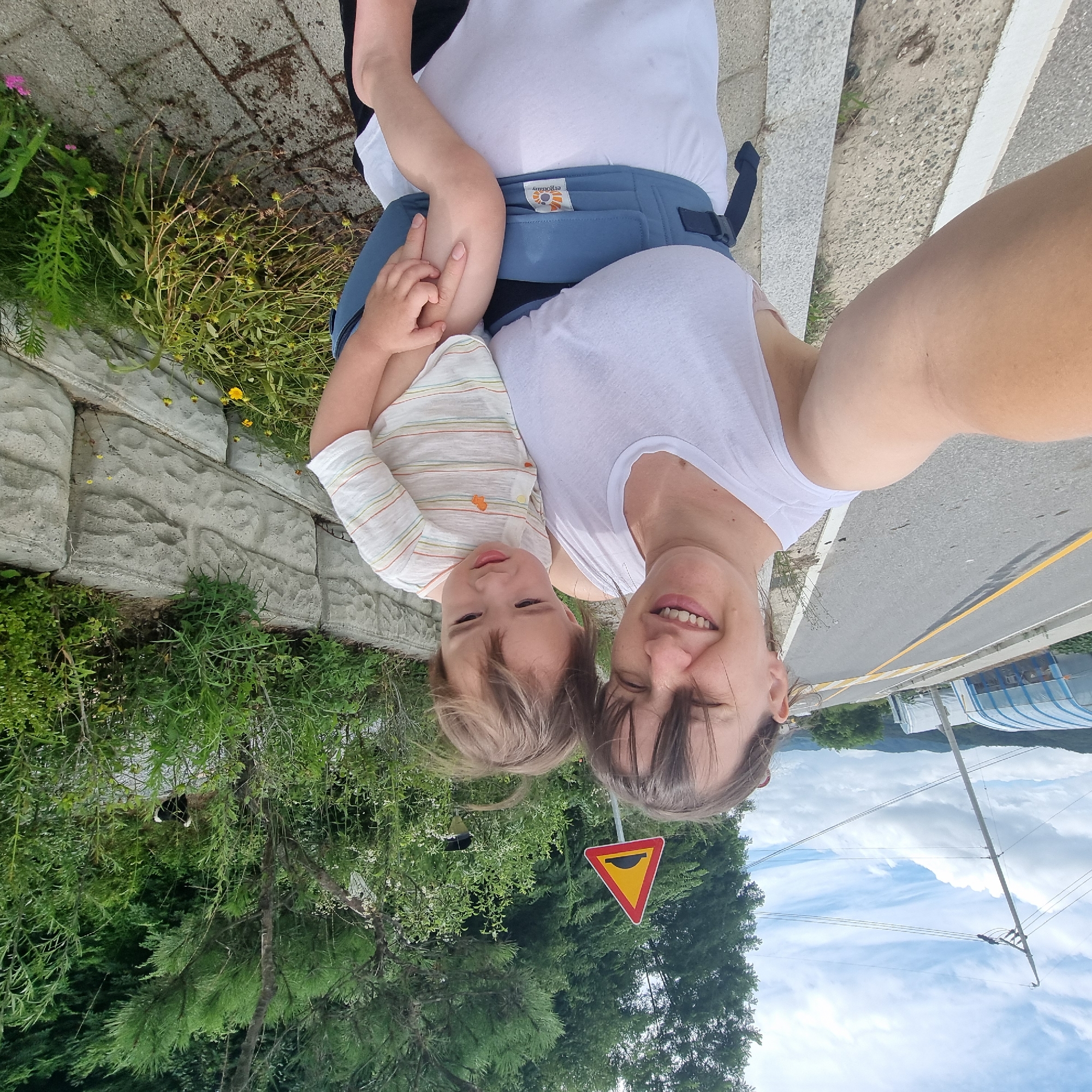 A woman is taking a selfie while holding a baby in a carrier. The woman is wearing a white tank top and has dark hair. The baby is wearing a striped shirt and has light brown hair. They are standing on the side of a road in Dunnae-myeon, South Korea. In the background, there is a speed bump sign, green trees, and some agricultural greenhouses. The road has a yellow line marking the center. A concrete retaining wall is visible behind them, with vegetation growing on top and around it.