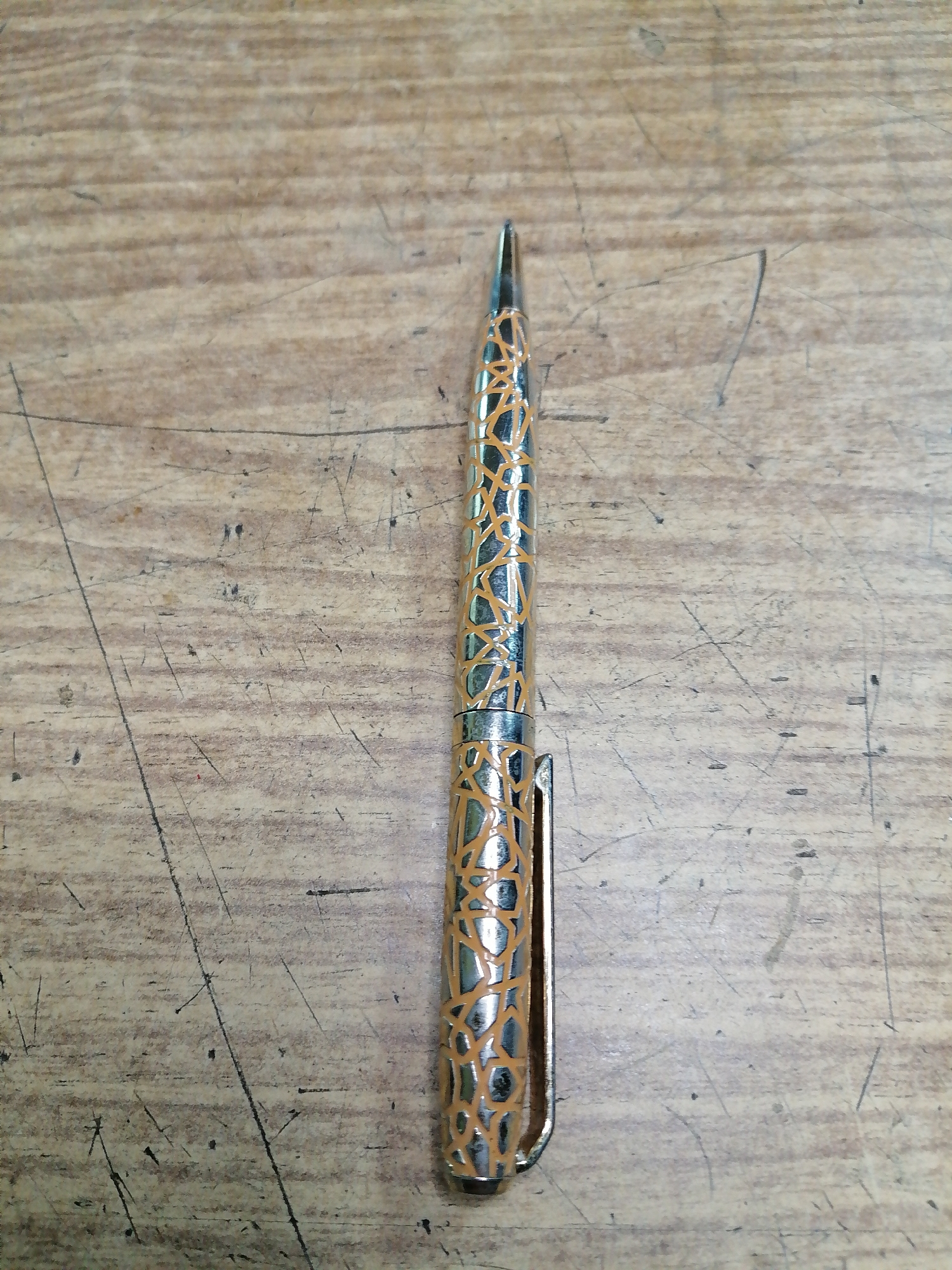 A single, silver and orange patterned pen is positioned vertically in the center of the image. The pen has a metallic sheen and a unique, abstract pattern of interwoven orange lines against a silver background. A silver clip is attached to the upper portion of the pen, extending slightly to the right. The pen rests on a wooden surface that appears to be a table or desk. The wood has a pronounced grain and is marked with numerous faint scratches, some of which are darker than others, suggesting wear and use. The lighting is even, casting no significant shadows. The overall impression is that of a still life, with the pen as the sole subject of focus. The image does not provide any information about the location in Rostov-on-Don, Russia.