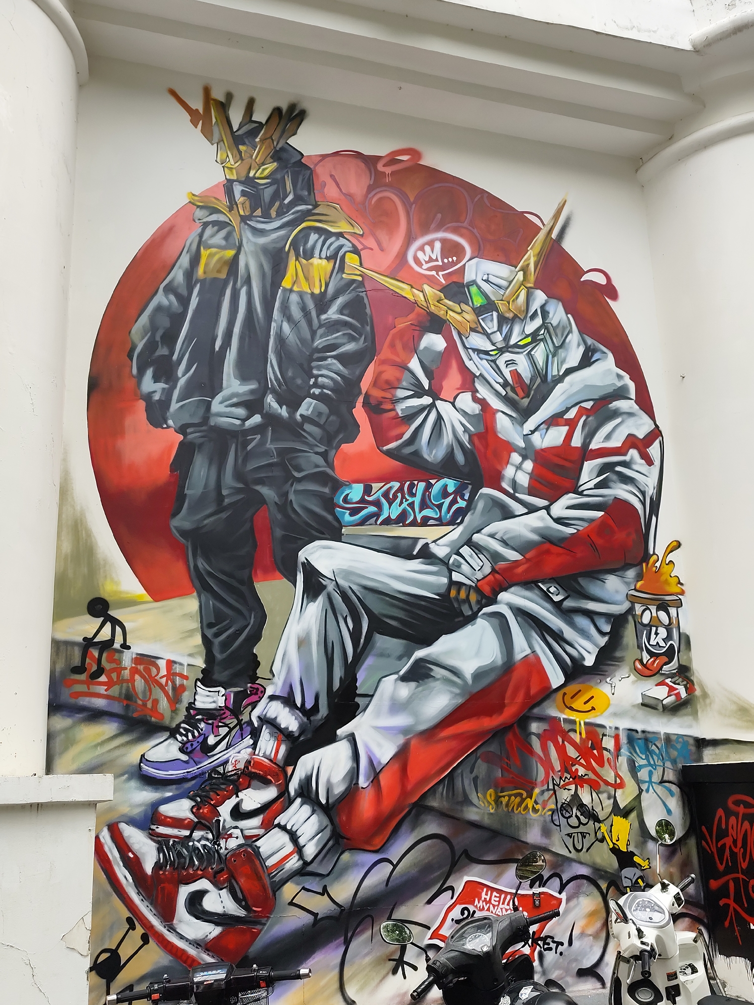 A mural decorates an exterior wall in Da Lat, Vietnam. The artwork features two anime-style figures in an urban setting. One figure stands, wearing a dark-colored jacket and pants, while the second figure sits, sporting a red and white hoodie and pants. Both figures have elements of robotic or mechanical design, notably in their headgear. The background is a red circle, with surrounding graffiti, including a coffee cup character. Sneakers adorn the figures' feet. Graffiti art covers the lower portion of the wall, blending with the mural. Two parked motorbikes rest at the bottom edge of the wall, partially obscuring the lower part of the mural. The mural is on a white wall between two cylindrical architectural features.