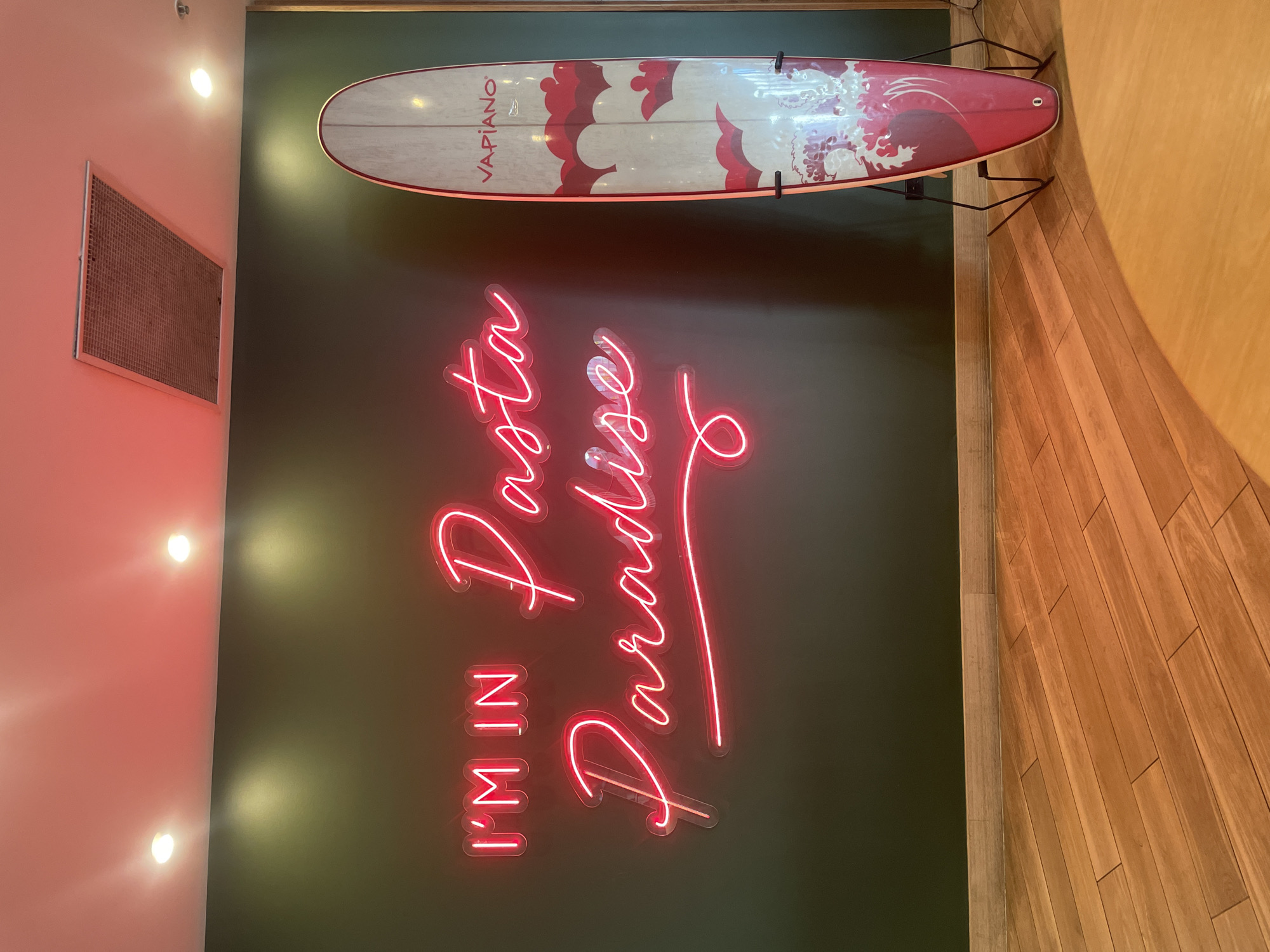 The image depicts an indoor scene with a dark green wall as the primary background. A neon sign on the wall displays the text "I'M IN Pasta Paradise" in bright pink. To the right of the sign, a surfboard stands upright on a black metal stand. The surfboard has a light-colored surface with a design of red and white clouds and waves. The text "VAPIANO" is visible in red at the top of the surfboard. The floor is made of light-colored wooden planks. The ceiling, illuminated by pink and white lights, has a rectangular vent. The setting does not specifically indicate the city of Surfers Paradise, Australia.