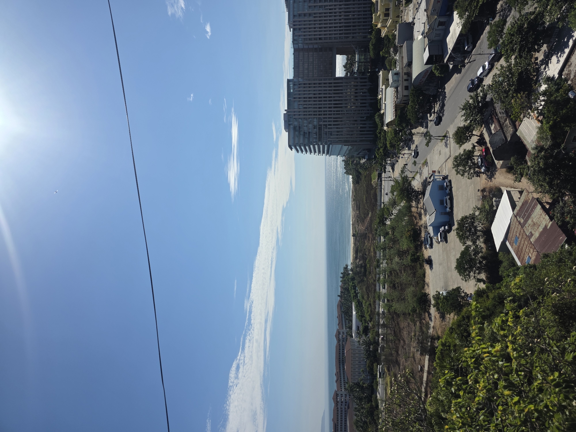 The image presents a wide panoramic view of a vibrant coastal city, likely Đà Nẵng, Vietnam, under a bright, sunny sky, captured from an elevated vantage point. The upper half of the frame is dominated by a clear, azure sky dotted with sparse, wispy white clouds. A brilliant sun flare is prominent in the top-left corner, and a small airplane is visible flying in the distance nearby. A thin, dark cable or power line extends horizontally across the upper part of the sky.
Below the sky, the glistening blue ocean stretches towards the distant horizon, meeting a long, sandy beach that borders the city. Along the coastline and further inland, a mix of architectural styles is visible. Modern high-rise buildings, some with visible balconies, stand alongside smaller structures. One distinctively large building features a prominent square cut-out or archway on an upper floor.
Interspersed among the buildings are roads, where several cars can be seen parked or driving, indicating normal daily activity. Lush green trees and vegetation cover various parts of the landscape, particularly in the foreground and on elevated terrain, suggesting the city is nestled against hills or a natural incline. The scene depicts a clear, bright daytime with excellent visibility, showcasing a lively urban and natural environment. No legible text is visible within the image.