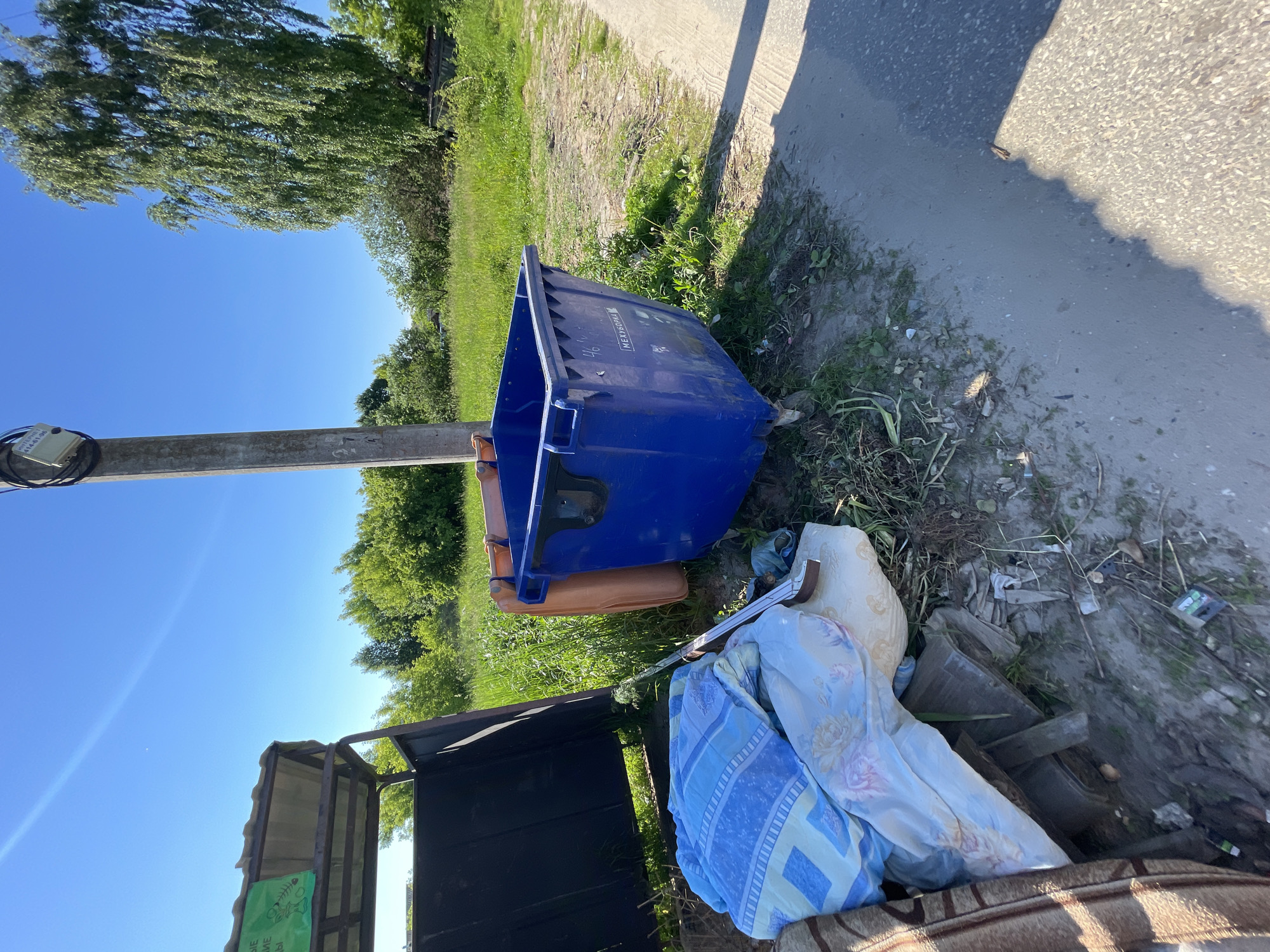 A blue industrial dumpster sits on a dirt and gravel shoulder next to a paved road in the village of Krasnye Baki, Russia. The dumpster is open and empty. To the left of the dumpster is a large pile of domestic refuse including blankets, pillows, and what appears to be discarded furniture. Behind the refuse pile and dumpster is a metal-framed structure with a sloped roof, covered on the left side with a black tarp. The area is bordered by a grassy field with trees in the background. A wooden utility pole stands to the right of the dumpster, with wires running from it. The sky is clear and blue.