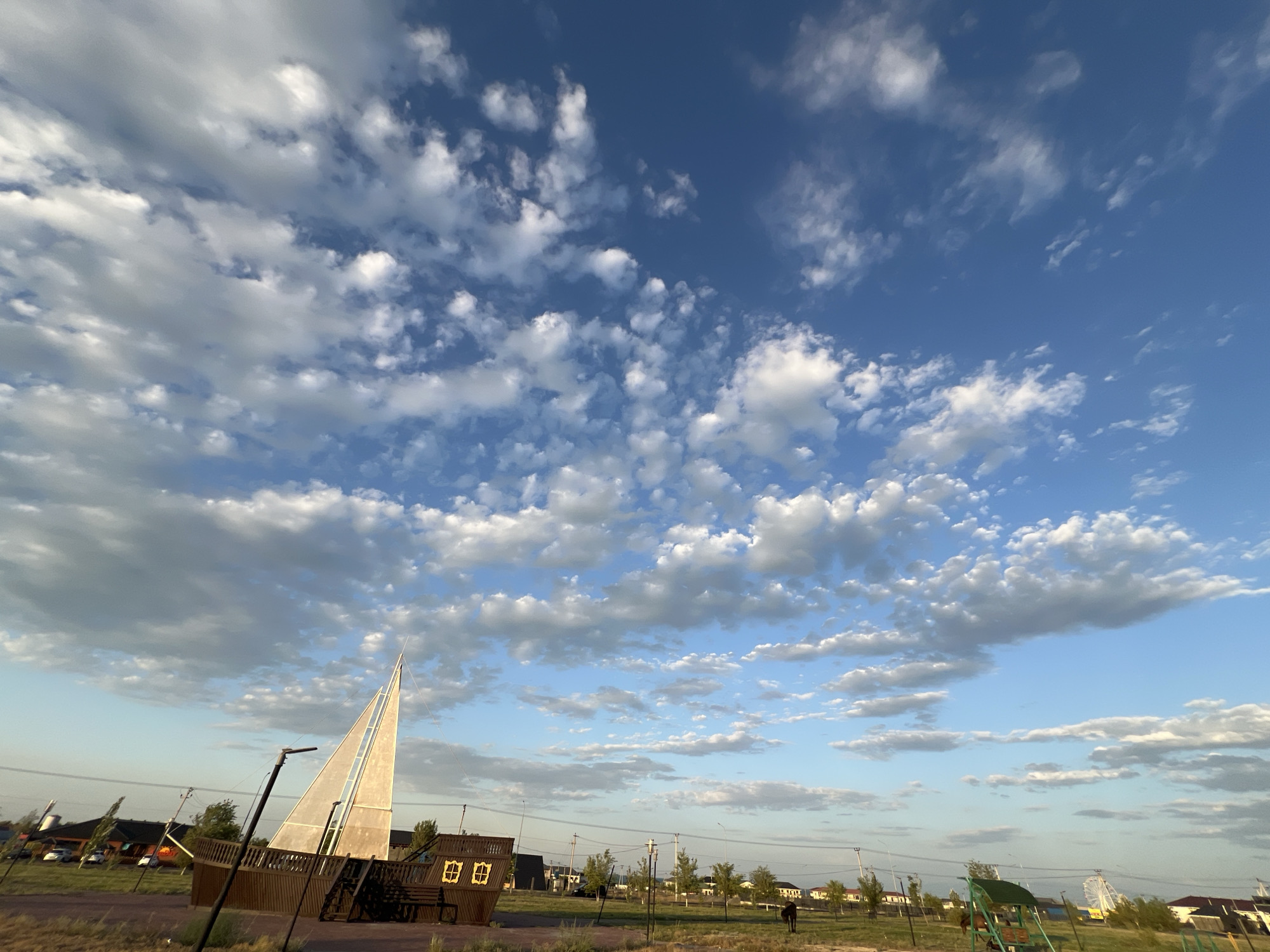 The image captures a park scene in the city of Ақши, Kazakhstan, under a vast blue sky filled with scattered white clouds. In the foreground, a wooden playground structure resembling a ship's hull with a tall, triangular white sail is prominent on the left. A dark lamppost stands in front of the ship structure. The ground appears to be covered in dry grass and dirt. In the middle ground, a line of trees separates the park from a residential area with low-rise buildings. Further into the background, the landscape stretches out with more trees and buildings. A few distant figures and what appears to be a horse can be observed in the park, and a Ferris wheel is visible on the far right horizon. The lighting suggests it is late afternoon or early evening, with soft shadows cast across the scene.