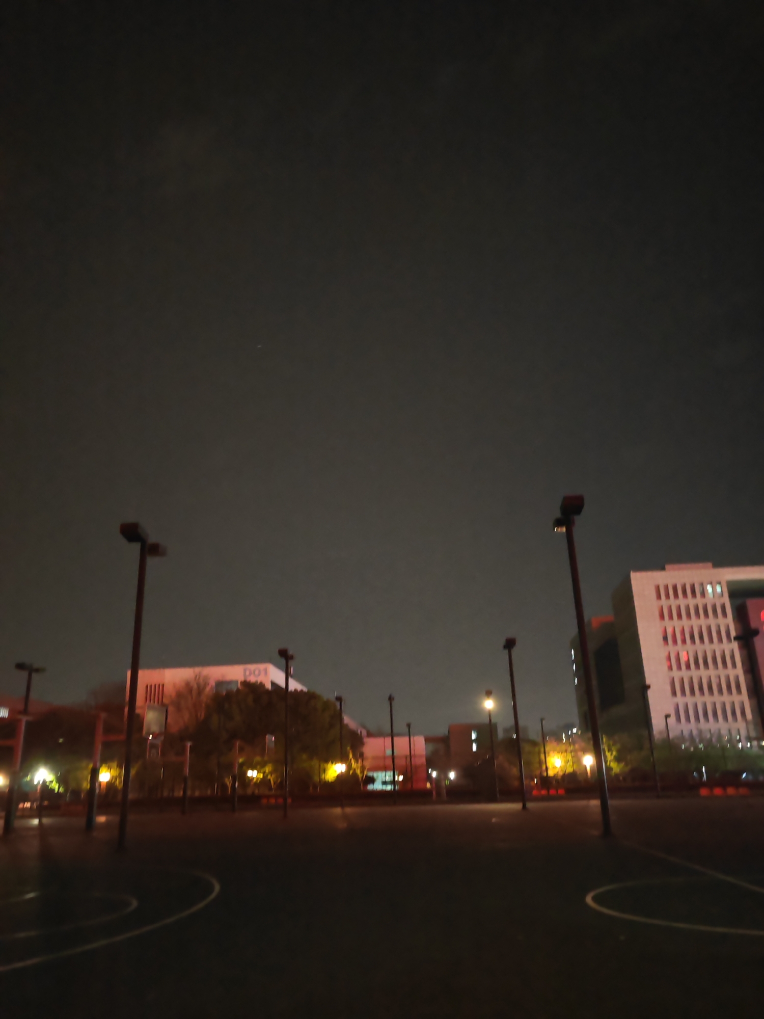 The image depicts an outdoor basketball court located in Jiangning District, China at night. The court surface is dark and reflective, and several light poles are positioned around it. Buildings are visible in the background, illuminated with artificial light. The sky is dark with minimal cloud cover. There are no people visible in the image.
