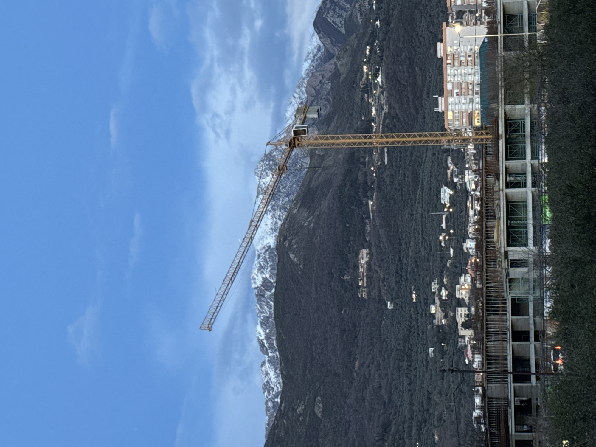 The image depicts a construction site in the city of Bar, Montenegro, against a mountainous backdrop under a partly cloudy, light blue sky. In the mid-ground, a tall yellow-orange tower crane stands prominently, its horizontal jib extending towards the upper left. Directly beneath and slightly in front of the crane is a multi-story concrete building under construction, showing exposed rebar and structural elements. The foreground is partially obscured by dense, dark green foliage.
Behind the construction elements, a vast, dark green, tree-covered mountain range rises steeply, its upper peaks distinctly snow-capped. Scattered across the mid-slopes of these mountains are numerous small clusters of buildings, many displaying illuminated windows, suggesting either early morning or evening. Between the construction site and the mountain's base, an urban or suburban area is visible, comprising various residential and commercial buildings, some with visible lighting. No individuals or specific actions are discernible within the frame.