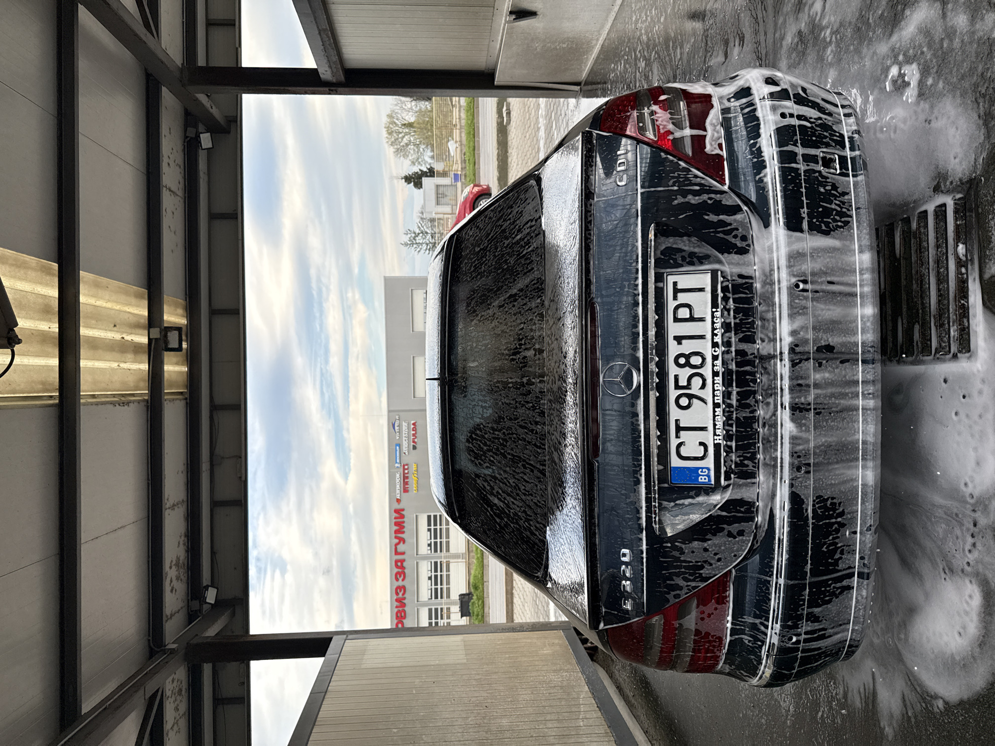 The image shows the rear of a black Mercedes-Benz E320 car covered in soap suds, presumably in a car wash. The car's Bulgarian license plate reads "CT 9581 PT". The text on the license plate frame states: "Нямам пари за G класа!" ("I have no money for a G class!"). In the background, there is a building with a sign that says "РВИЗ ЗА ГУМИ", with multiple tire brand logos including Pirelli, Goodyear, and Bridgestone. Based on this sign, the location is likely Stara Zagora, Bulgaria, as indicated in the request. The car wash structure is made of metal framing and light-colored panels. The sky is visible in the background, suggesting an outdoor location. The car is positioned on a grid-like platform, typical of car washes.