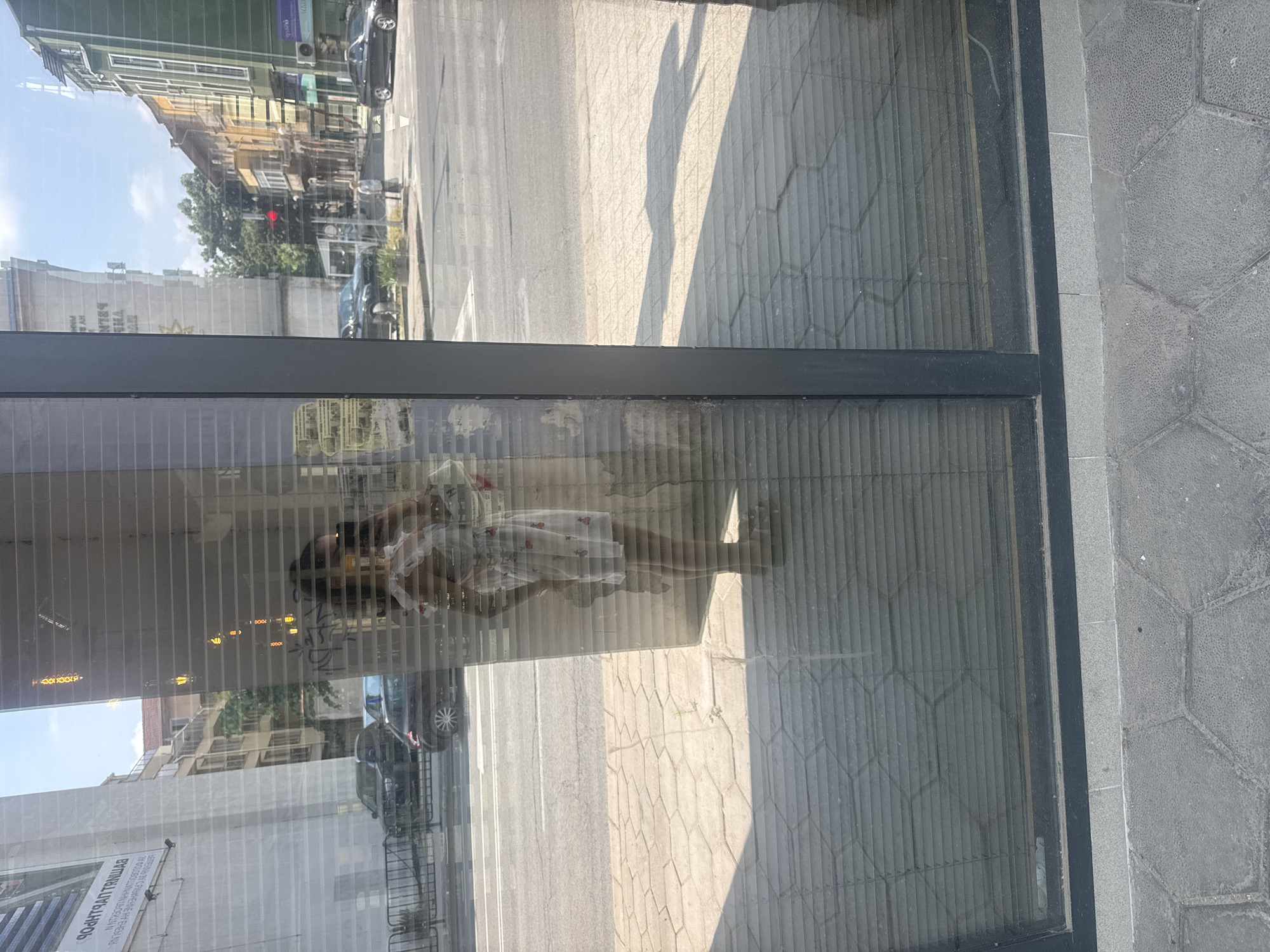 An outdoor shot captures a complex layered scene through a large glass window, reflecting the street and buildings, while also showing the interior through vertical blinds. The image is taken during the daytime under sunny conditions, casting distinct shadows on the ground.
In the foreground, a window frame separates the sidewalk from the building. The sidewalk is paved with grey hexagonal stones. Through the glass, a street or paved area is visible, with sunlight illuminating patches of it. In the reflection, a person, likely a woman, is seen wearing a light-colored dress with a red pattern, possibly holding an object near her face or shoulder. She appears somewhat translucent due to the reflections.
The reflections also show various urban elements: multi-story buildings with different architectural styles, some with balconies, and green trees. Several cars, including a dark-colored sedan, are parked or driving on the street. A red traffic light is faintly visible amidst the greenery, and a pedestrian crossing sign is also reflected.
Behind the reflections, the vertical blinds of the building's interior are clearly visible, creating a striped pattern across the glass. On a large white sign or banner reflected on one of the buildings, reversed Cyrillic text can be partially read as "УРРАТИЯ" (URRATIA), among other indistinguishable characters. The overall impression is a bustling urban environment viewed through a reflective surface, blending interior and exterior elements.