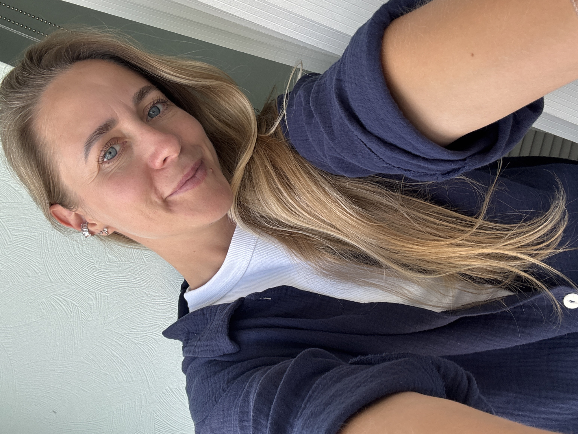 This is a selfie of a woman with blonde hair and blue eyes, wearing a navy blue button-up shirt over a white t-shirt. She is wearing earrings in her left ear. The background appears to be an interior or exterior wall with a textured pattern. There is no identifiable information within the image that places the location in Chaikovsky, Russia.