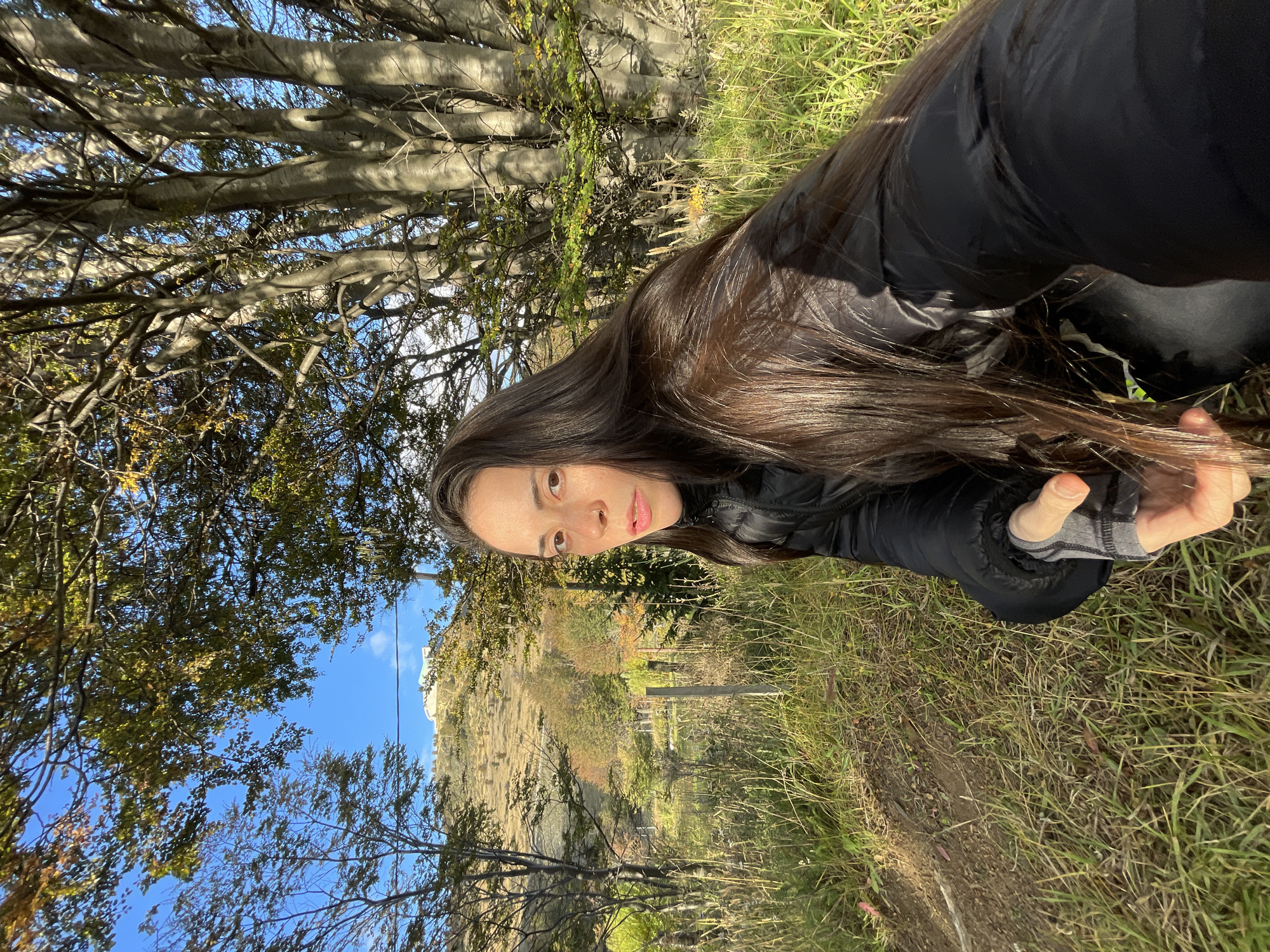 A woman with long, dark hair wearing a black puffer jacket is positioned in the foreground, looking at the camera. She's holding up her hand, giving a thumbs-up gesture. The background reveals a landscape typical of Ushuaia, Argentina. The setting includes trees, grassy areas, and a hill or cliff. The sky is partially visible through the trees, showing blue with some clouds. A building is visible on the hill in the background. The image is taken outdoors, with natural light.