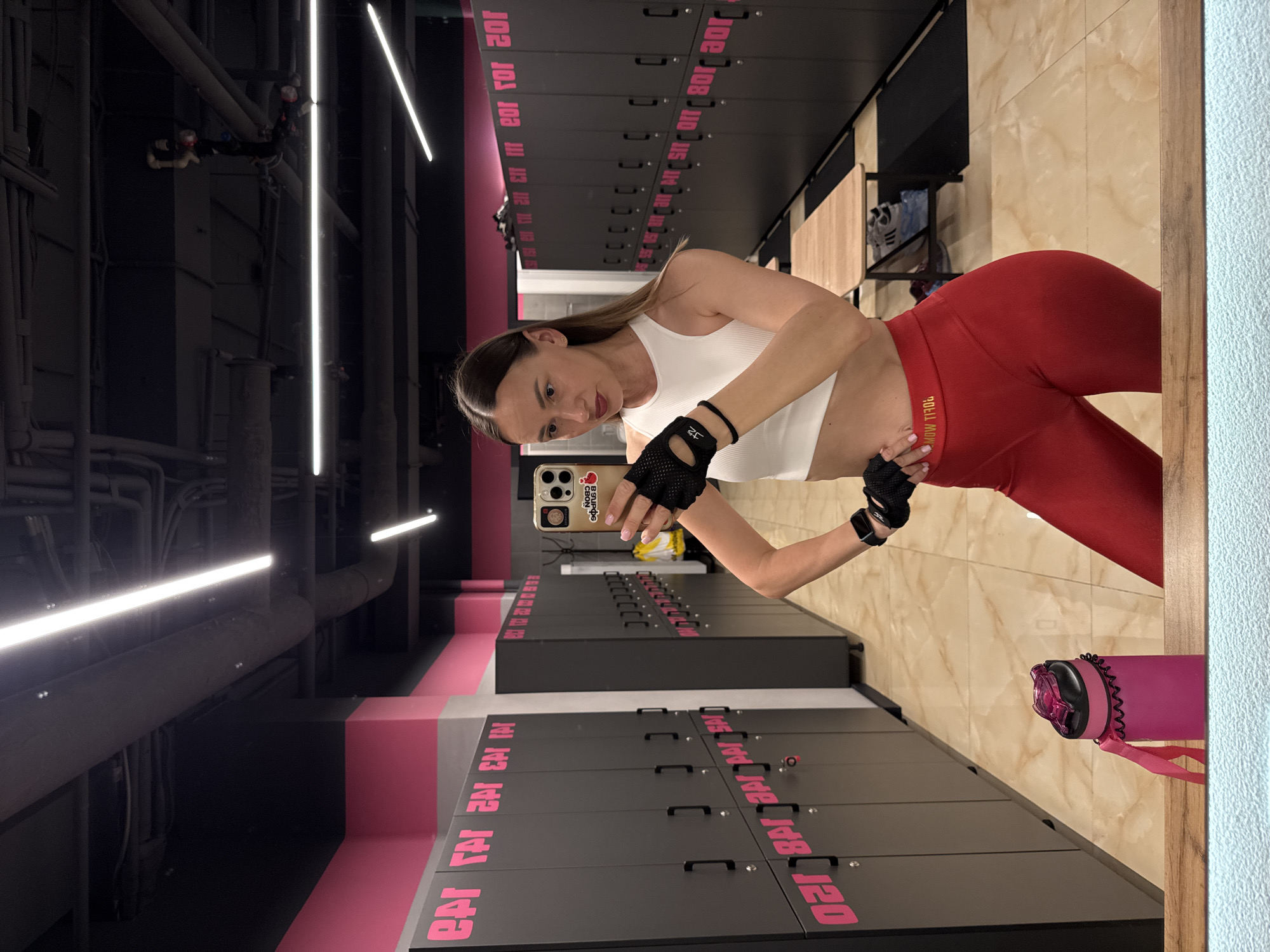 A woman is captured in a mirror selfie within what appears to be a modern gym or locker room setting in Sochi, Russia. She has long, dark hair and is wearing a white sports crop top, red leggings, and black fingerless gym gloves. She holds a light-colored smartphone with a case featuring "B SUPOS CBON" text and a small red design, angled to take the picture of her reflection. Her left hand rests on her hip, accentuating her pose. The waistband of her red leggings displays "BOLIL MON" in gold text.
The background shows rows of dark gray lockers with prominent pink numbering and lettering. Visible locker numbers include "102," "103," "108," "140," "141," "142," and "143." Some lockers also bear stylized pink text that repeatedly resembles "EAT." Wooden benches are positioned in front of these lockers. The ceiling reveals an industrial aesthetic with exposed pipes and bright, linear LED strip lights. A pink water bottle with a dark lid is visible on the floor to the right. The space is well-lit, suggesting an indoor daytime or brightly lit environment. The woman's expression is neutral and composed for the photograph.