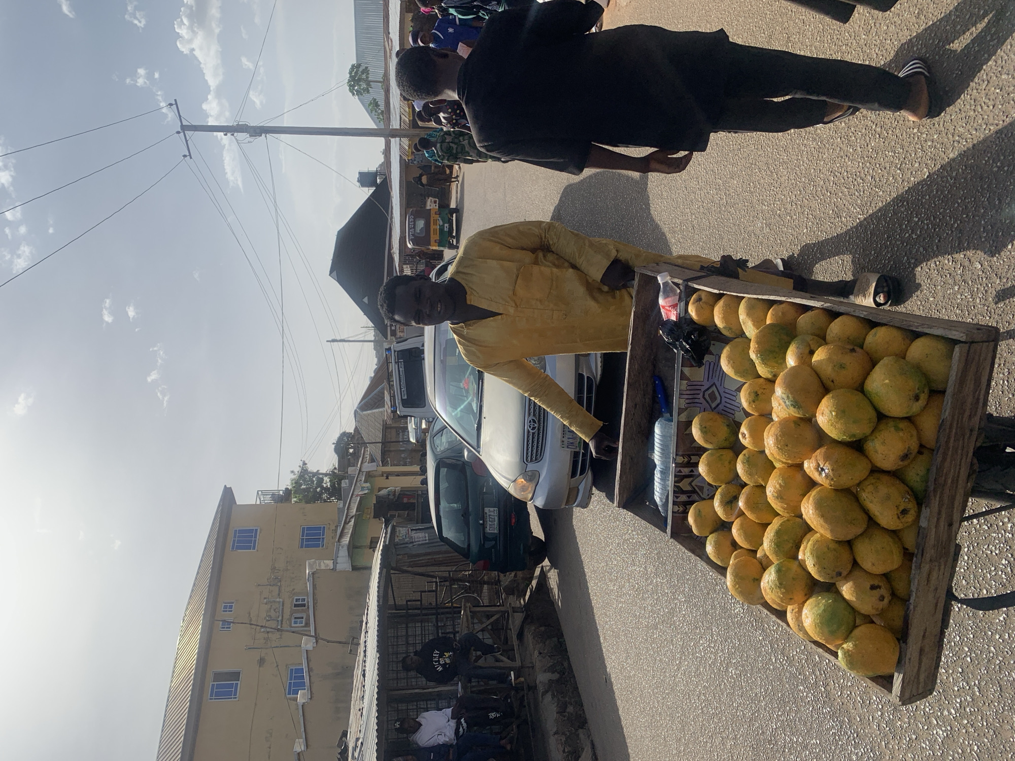 On a street in Jos, Nigeria, a man in a yellow shirt stands beside a cart filled with mangoes. A second person walks past, and cars are parked along the street. Buildings are visible in the background.
