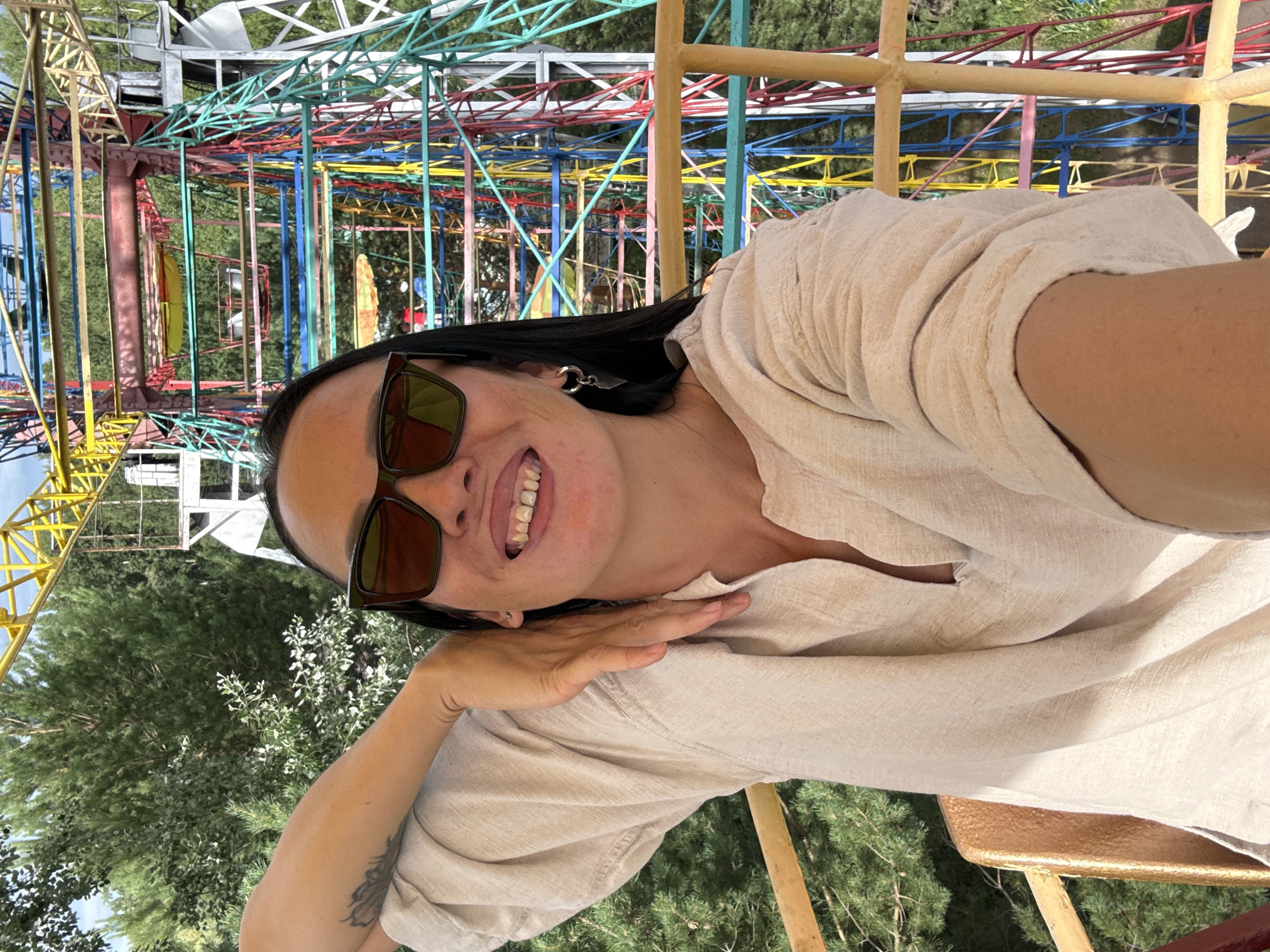 A woman with dark hair and sunglasses is seated in a gondola of a Ferris wheel, smiling with her teeth showing. She is wearing a light-colored, loose-fitting shirt and a silver hoop earring. Her left arm is bent with her hand resting on her neck. Behind her, the colorful structure of the Ferris wheel is visible, along with green trees and a bright sky. The Ferris wheel is located in the city of Chaikovsky, Russia.