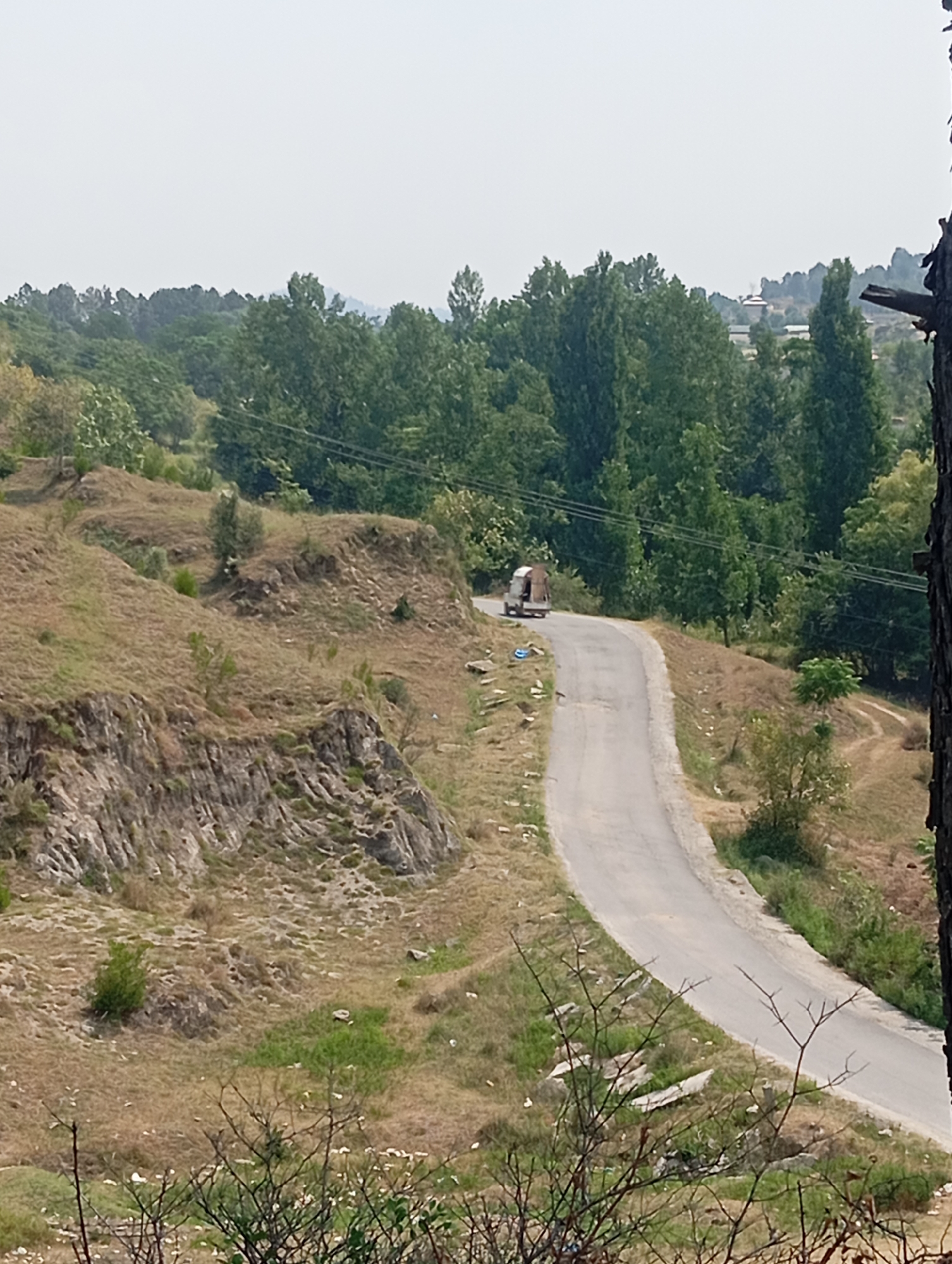 A winding, paved road curves through a hilly, sparsely vegetated landscape. The road is bordered by dry, grassy slopes with rocky outcroppings on the left and a denser line of trees in the distance on the right. A small, utilitarian vehicle, possibly a three-wheeled van, is traveling away from the camera on the road. The sky is hazy and overcast. The scene appears to be in a rural or semi-rural area, consistent with the outskirts of Hakale, Pakistan.