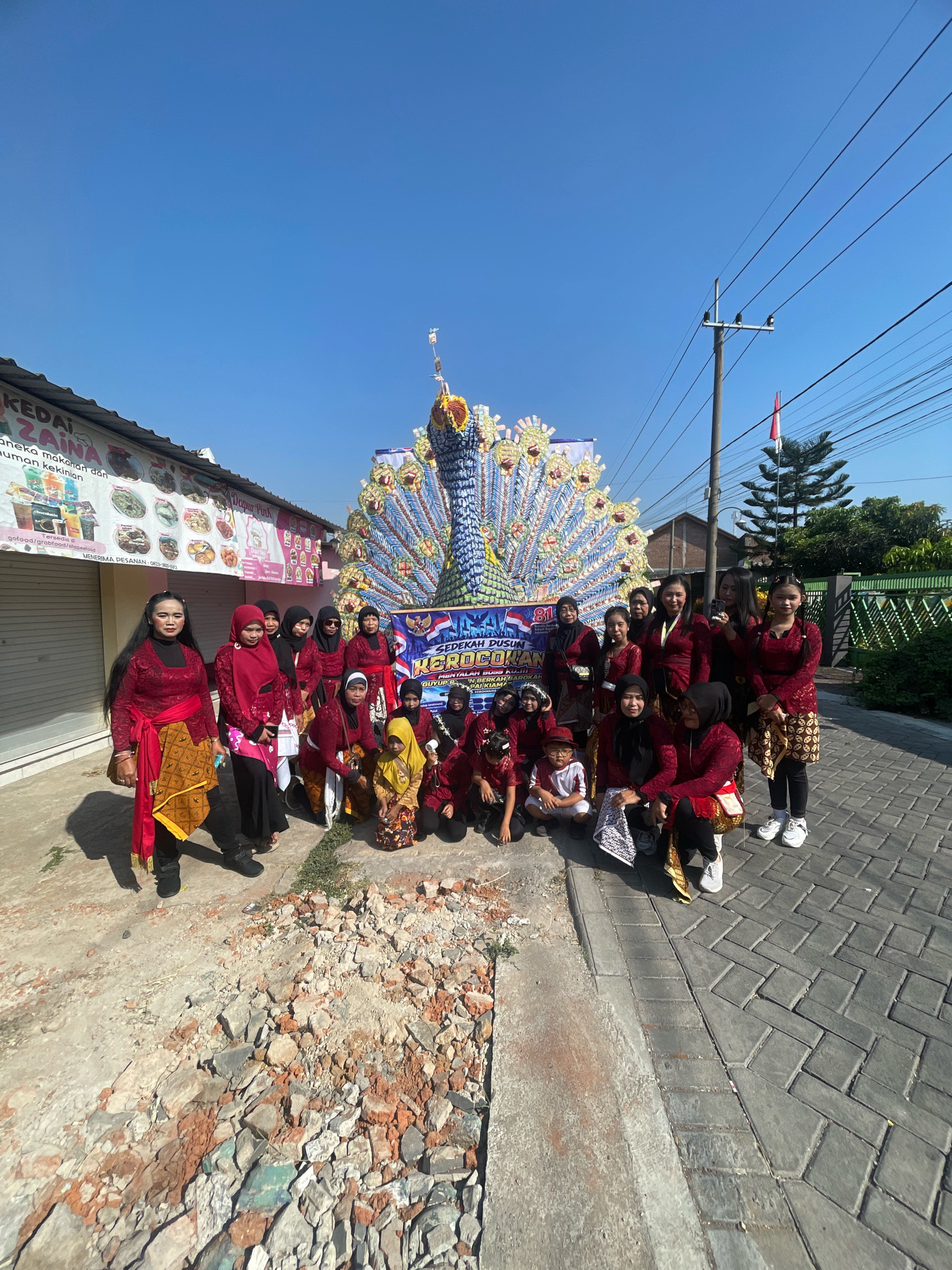 A group of women and children are posing in front of a large, colorful peacock float on a sunny day. They are wearing traditional Indonesian attire, primarily in shades of red, maroon, and gold. The peacock float is elaborate, with a blue body and a fanned tail made of many small, colorful elements, possibly plastic bottles or similar recycled materials. A banner with text in Indonesian, including "SEDERAH DINUSAN GROKON," is visible behind the float. To the left of the float is a building with a shopfront displaying various food items and products. The ground in front of the building is a mix of uneven dirt, scattered bricks, and a paved walkway made of interlocking gray bricks. In the background, there are power lines, a wooden utility pole with insulators, and a tall green pine tree. The sky is clear and bright blue. The scene appears to be outdoors in a public area, likely a street or a gathering place in Kebon Waris, Indonesia, given the context provided.