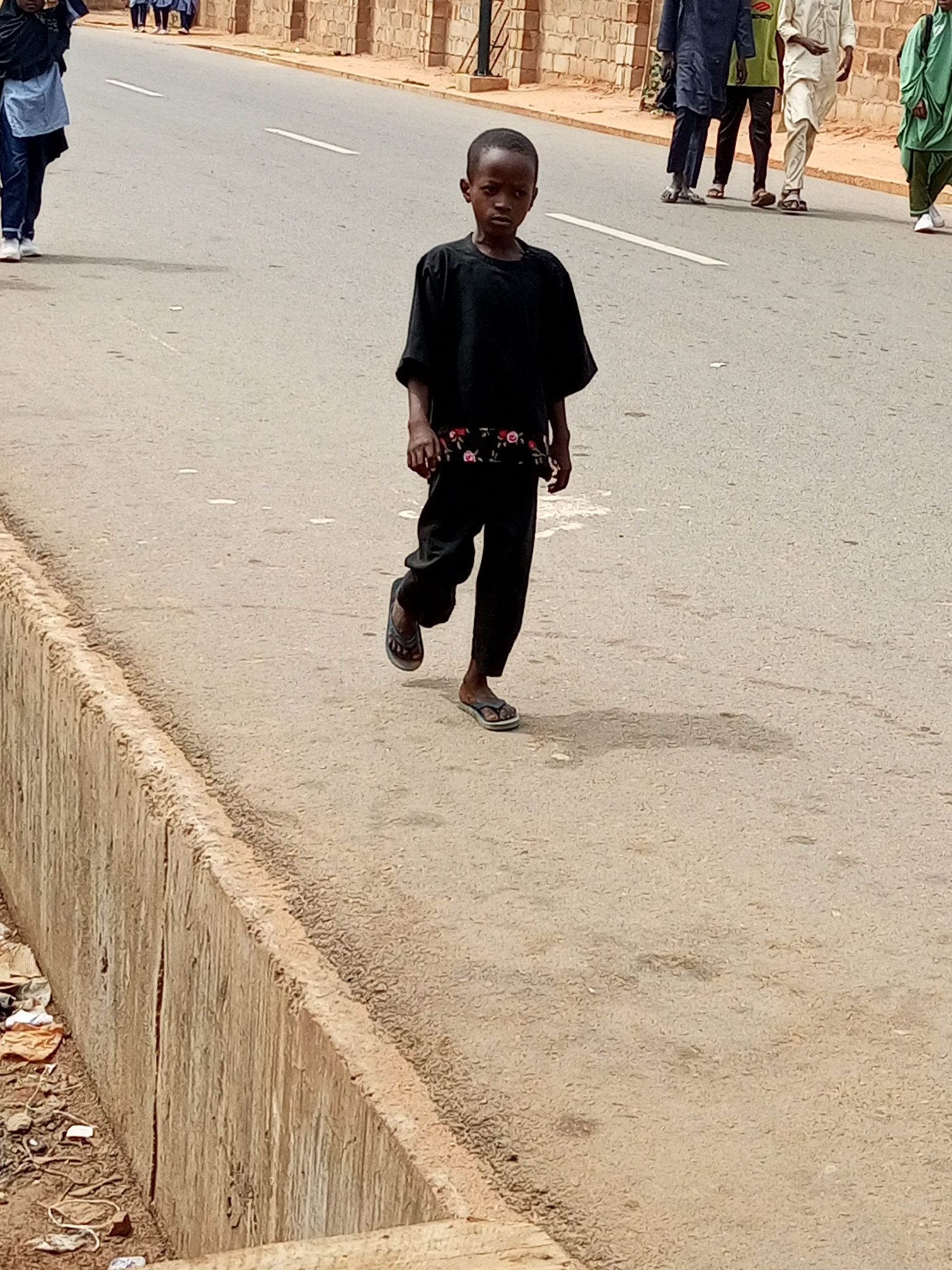 The image shows a young boy in a black t-shirt and black pants walking on a paved street in Gombe, Nigeria. The boy is in the foreground, looking towards the camera. Behind him, on the street, several adults are walking away from the camera. They are wearing various traditional Nigerian outfits. The street has white lines painted on it. To the left of the street, there is a concrete embankment with debris at its base. The background features a brick wall and buildings. The overall setting appears to be a public street in an urban environment.