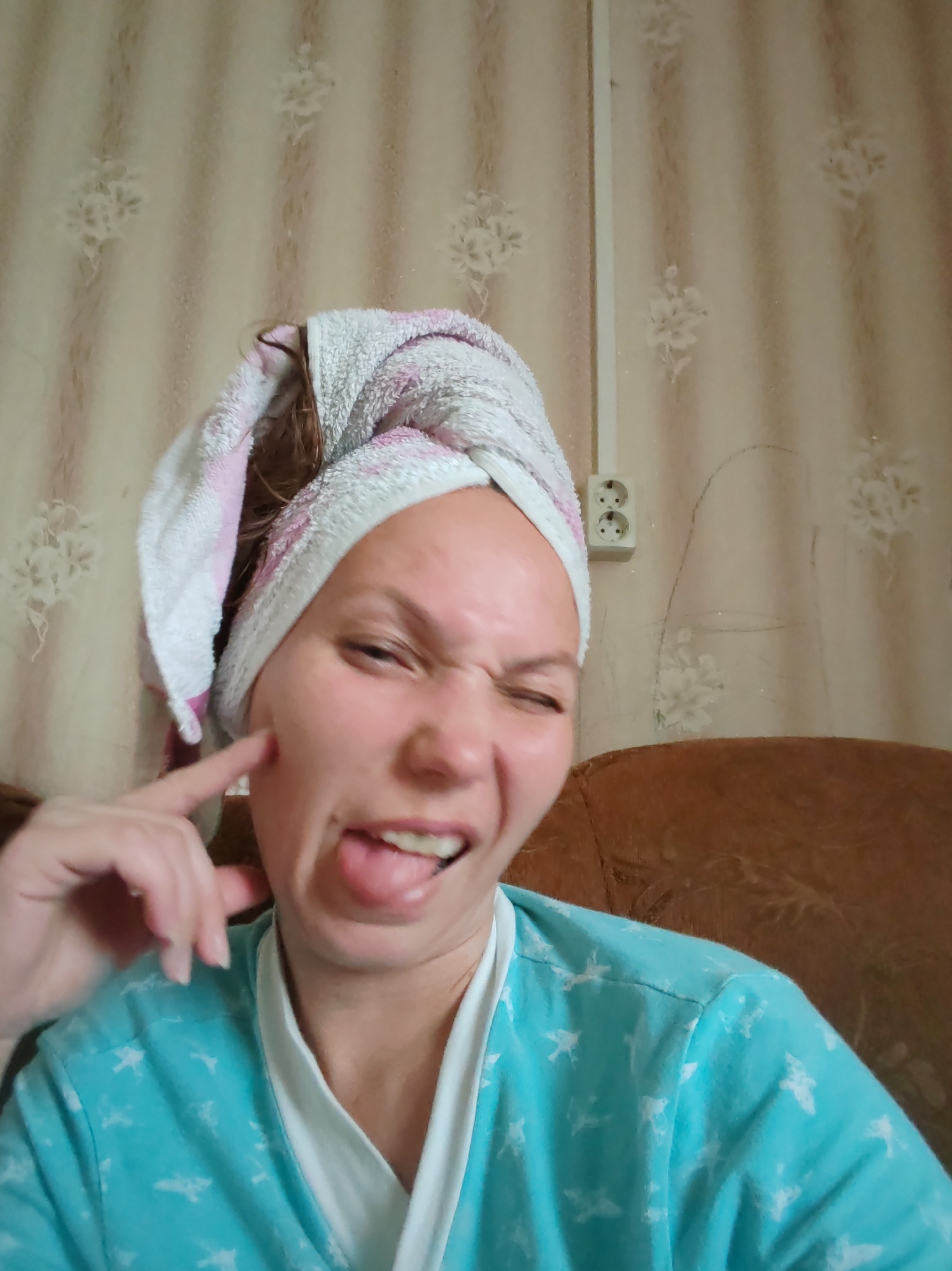 The image shows a woman with a towel wrapped around her head and wearing a blue robe with a white trim. She is making a funny face, with her tongue sticking out and one eye squinted. Her left hand is raised to her cheek. The background appears to be a room with patterned wallpaper and a brown upholstered chair. There is an electrical outlet on the wall. The overall impression is one of a casual, playful moment. The location is identified as Чухлэм, Russia.