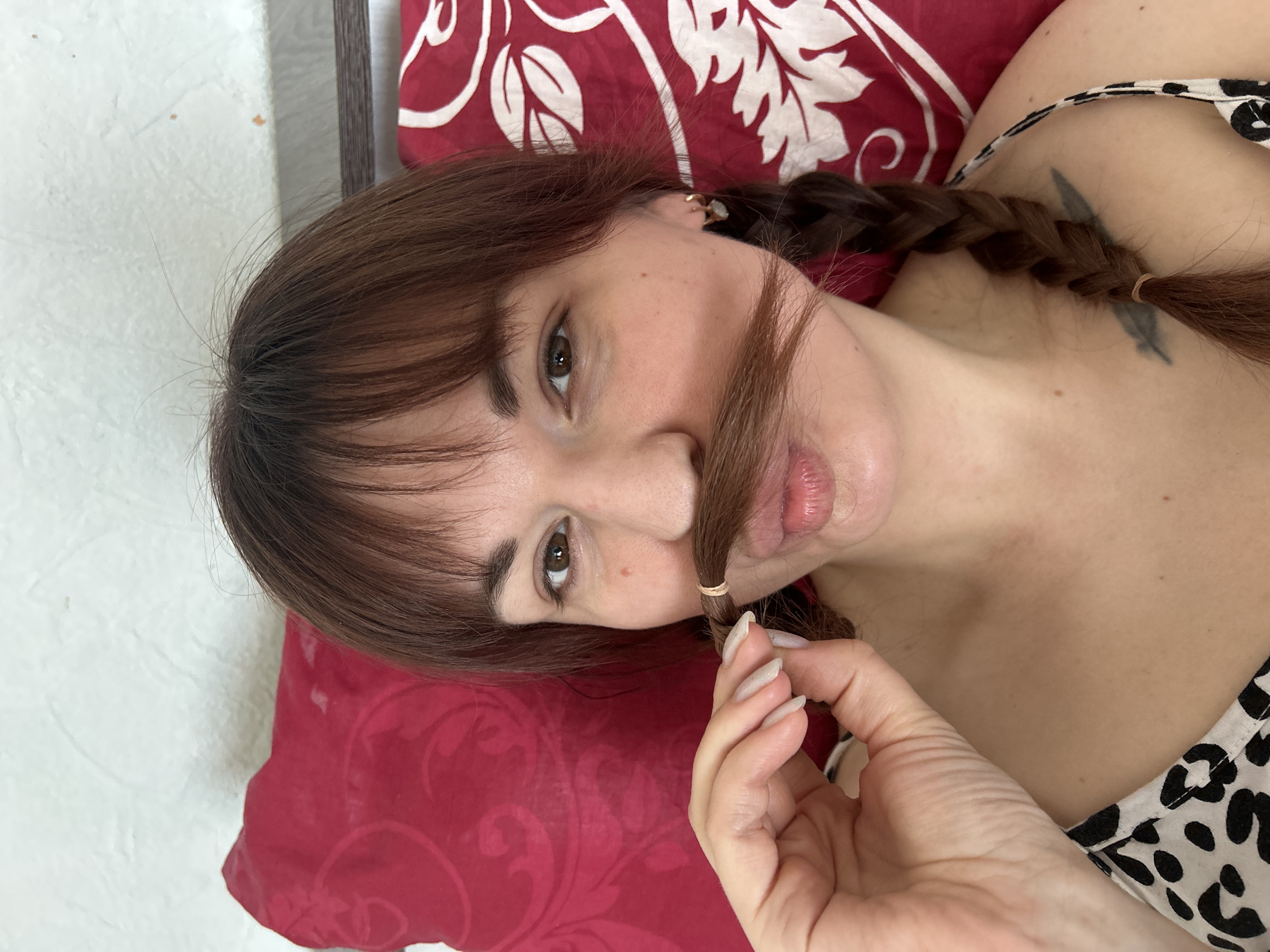 In the city of Фоки, Russia, a close-up, head-and-shoulders portrait depicts a single individual, presumably female, lying or reclining. She possesses light brown hair styled with bangs, and her brown eyes are directed at the camera. A section of her hair, secured with a light brown elastic band, is positioned above her upper lip, simulating a mustache. Her right hand, with manicured, light-colored fingernails, holds this hair segment in place. Her lips are slightly pursed. Her hair is braided into two pigtails, with one braid visible resting on her right shoulder, secured at the end with a light brown elastic band. She is wearing a top with a black and white leopard print strap visible on her right shoulder. Small, gold-colored hoop earrings are visible in her right ear. A small, raised brown mark is present on her right cheek. A dark, faded tattoo, appearing to be a feather or leaf shape, is visible on her right collarbone area, partially obscured by the braid. The background consists of a red pillow with a subtle, darker red floral pattern, behind which a textured, light-colored wall and a dark, narrow horizontal element are visible.