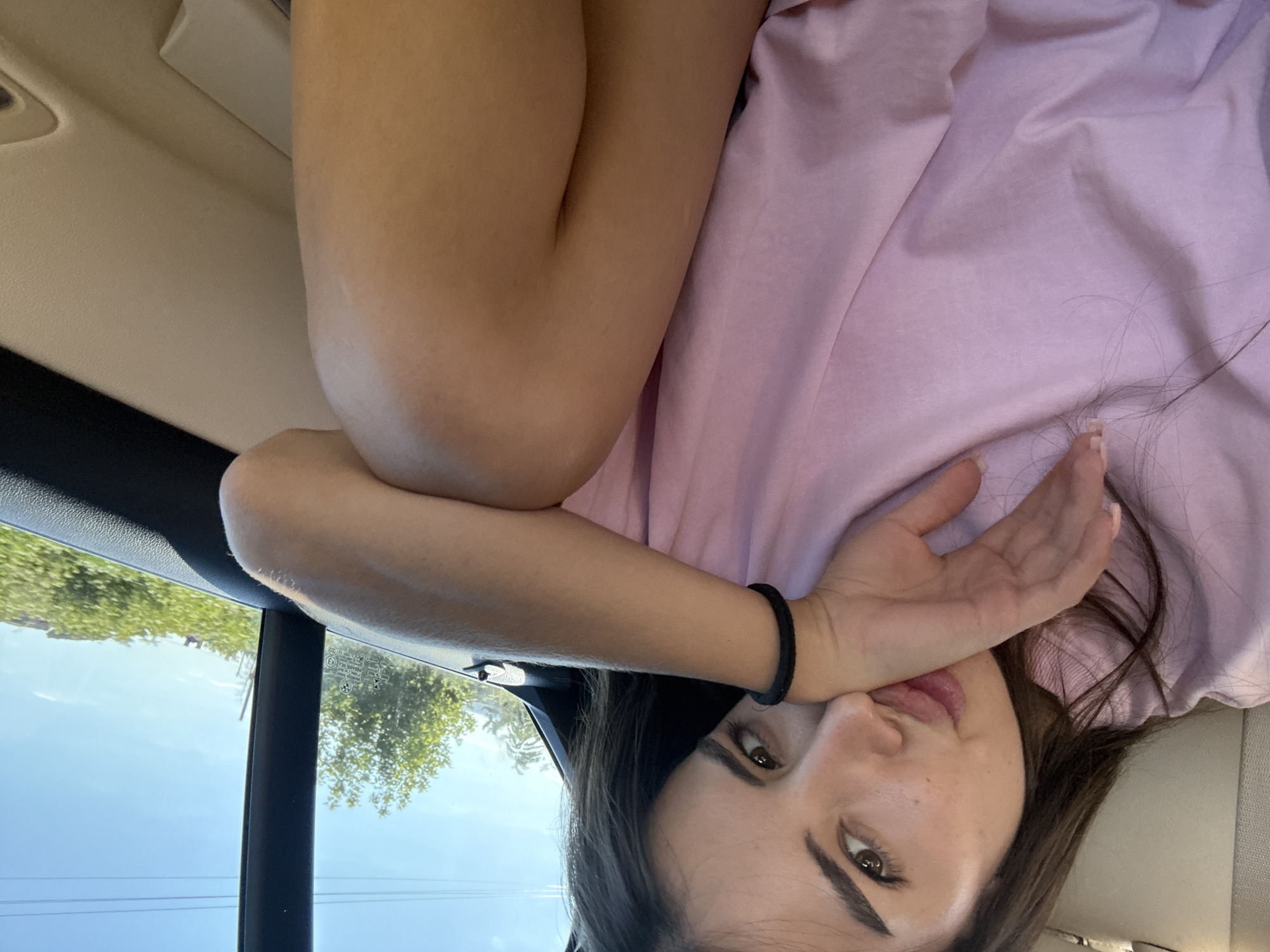 A young woman is seated in the back of a vehicle, looking towards the camera with her hand resting on her mouth. She is wearing a light pink t-shirt. Her eyes are brown, and her hair is dark brown. Through the window, a clear blue sky and green trees are visible, suggesting an outdoor environment. The interior of the car appears to be upholstered in a light-colored material.