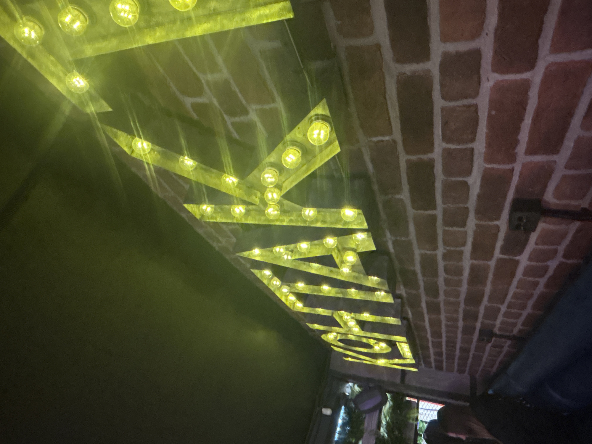 The image captures an interior space with a distinctive aesthetic. A decorative lighting fixture dominates the scene, shaped like the letter "Z" constructed from strips of material illuminated with numerous round light bulbs that cast a bright yellow glow. This fixture is mounted on a ceiling composed of red brickwork. The wall to the left of the lighting fixture is a dark, solid color, contrasting sharply with the brightness of the "Z" light. Below, visible through a window or opening, is a glimpse of foliage, possibly indicating an outdoor space or garden. Though further identifying details are unavailable, the location is specified as Saint Petersburg, Russia. However, the photograph focuses solely on the interior details.