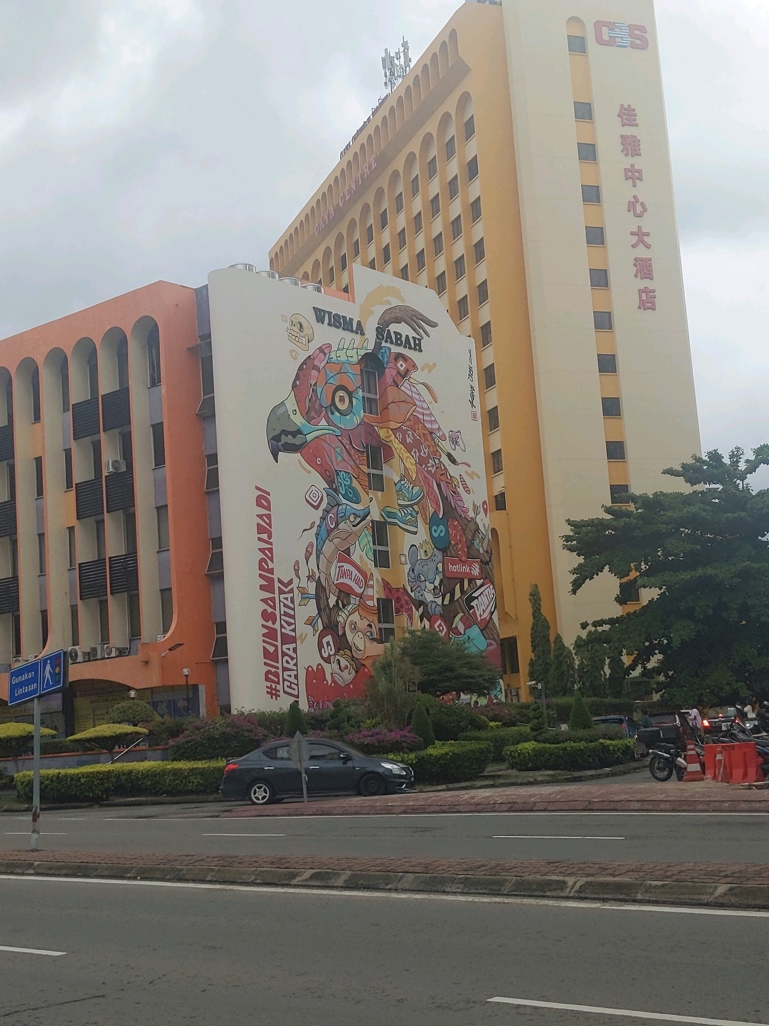 This image captures a street scene in Kota Kinabalu, Malaysia, focusing on two buildings and the surrounding environment. A tall, light-yellow building dominates the right side of the frame, featuring rows of dark rectangular windows and prominent signage at the top with the letters "CES" and vertical Chinese characters. Attached to this building on the left is a smaller, orange building with arched window openings and external air conditioning units visible.
A large, colorful mural covers a significant portion of the white wall between the two buildings. The mural depicts a stylized bird, possibly an eagle, surrounded by various cartoonish figures, text, and design elements. The text "#BIKINSAMPAIJADI" and "CARA KITA" are prominent in white letters against a red background.
In the foreground, a multi-lane road runs horizontally across the image, with a dark sedan parked near the curb on the left. A pedestrian crossing sign with a blue background and white figures indicating a pedestrian is visible to the left of the mural. Landscaping with manicured bushes and flowering plants lines the area between the road and the buildings. To the far right, several motorcycles and traffic cones suggest an area of pedestrian or vehicle activity. The sky is overcast, casting a diffused light over the scene.