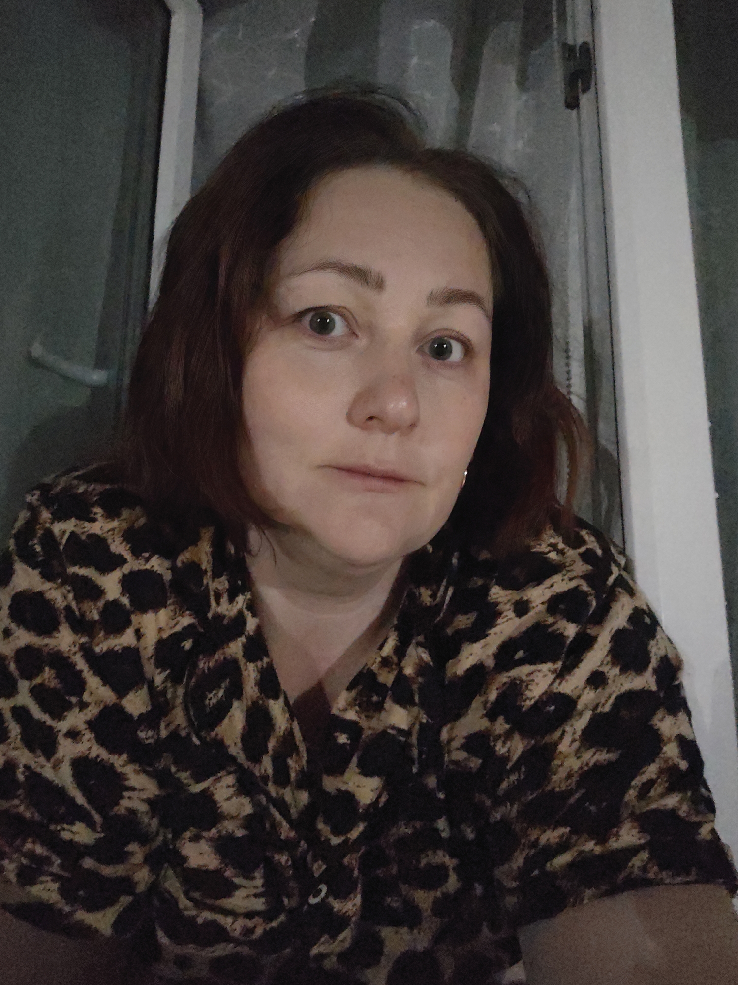 A woman is centered in a medium close-up shot. She has light skin, reddish-brown hair, and is wearing a leopard print shirt. Her facial expression is neutral. She appears to be indoors, possibly in a residential setting, with a window and wall visible in the background. The location is Markovskii, Russia. The image doesn't show activities or interactions but focus on presenting her within the given setting.